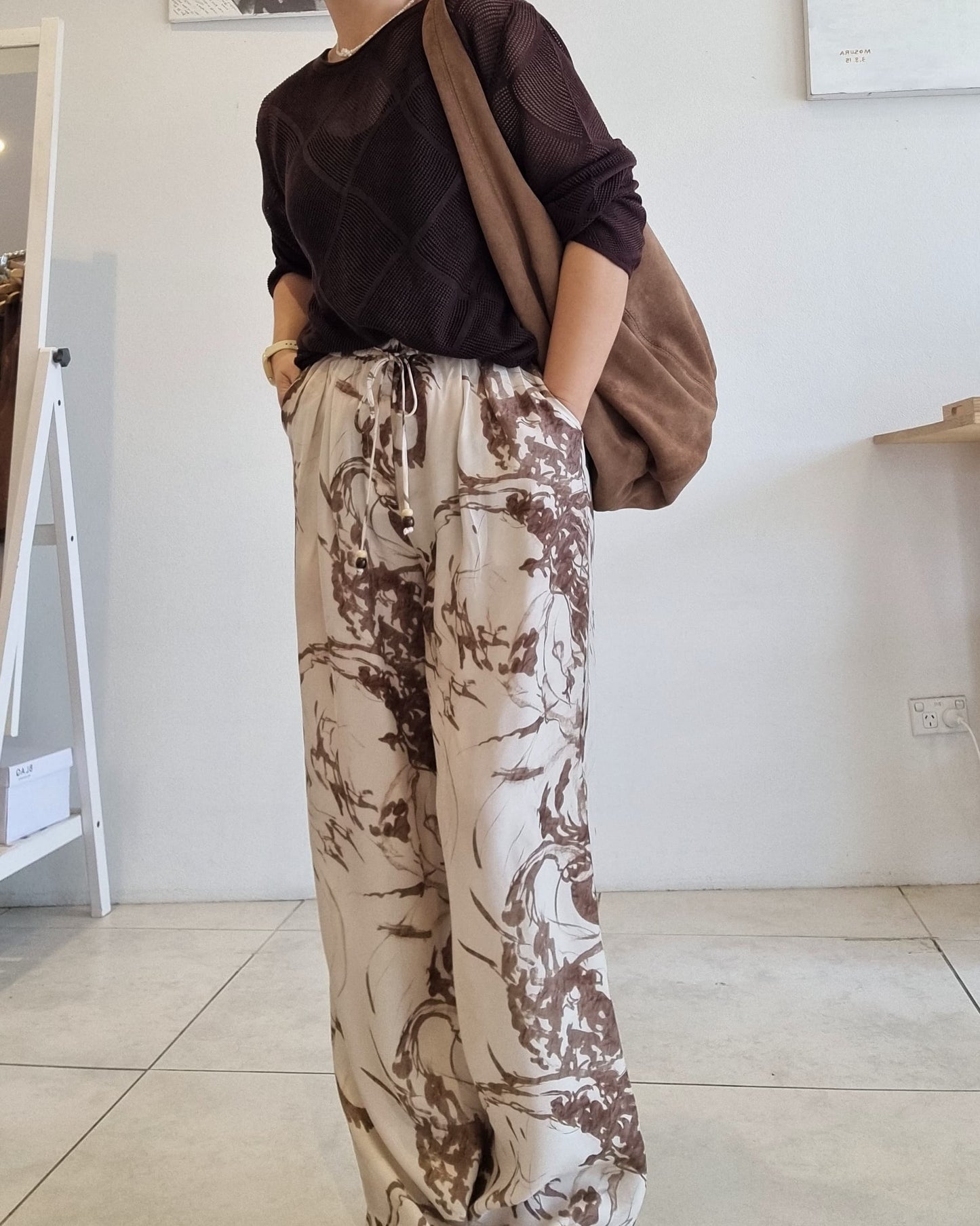 Side view of brown Tencel mesh knit long-sleeve top styled with beige abstract print wide-leg pants and a brown shoulder bag.