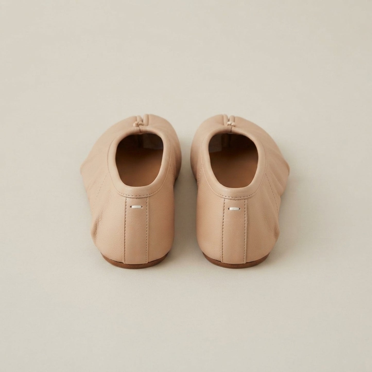 A pair of light beige leather ballet flats shown from the back on a neutral, light beige background.