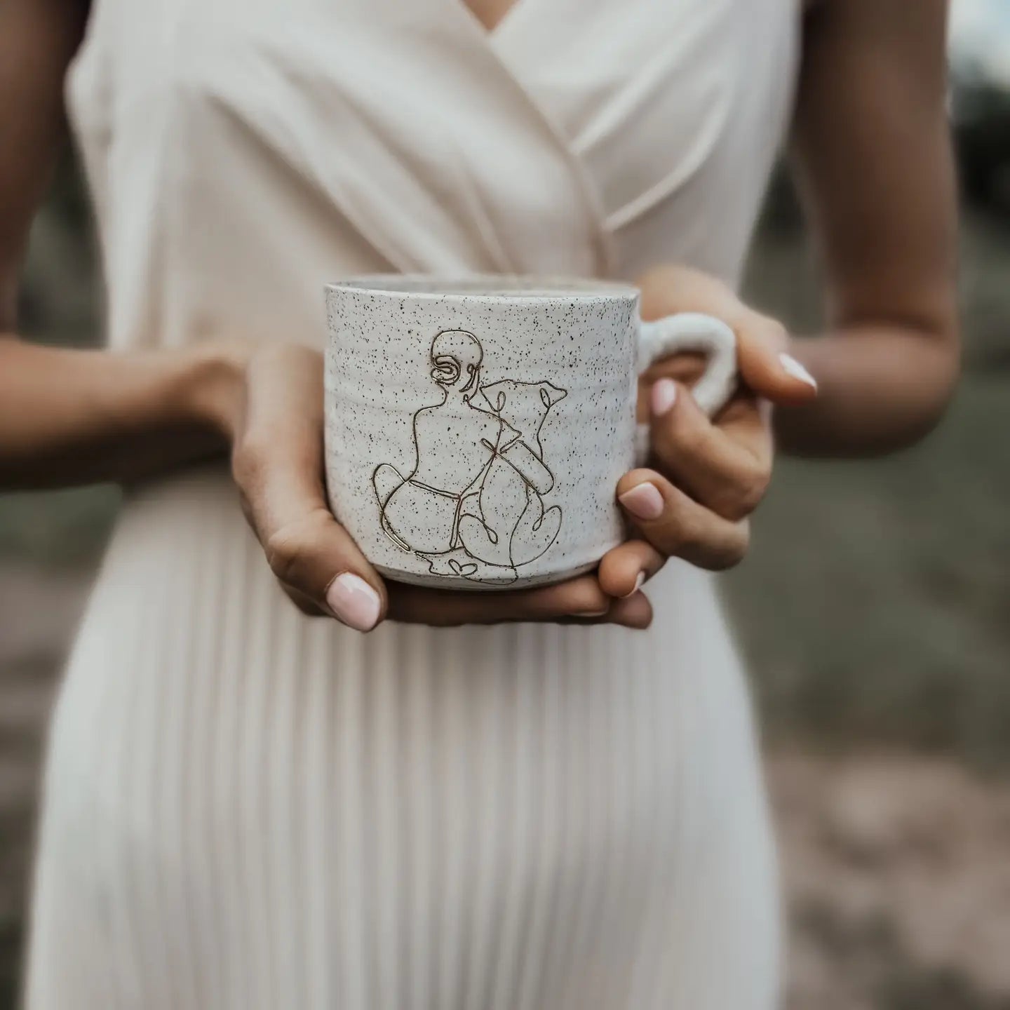 Woman in a cream dress holding a speckled ceramic mug featuring a minimalist girl-and-dog illustration, with a softly blurred outdoor background