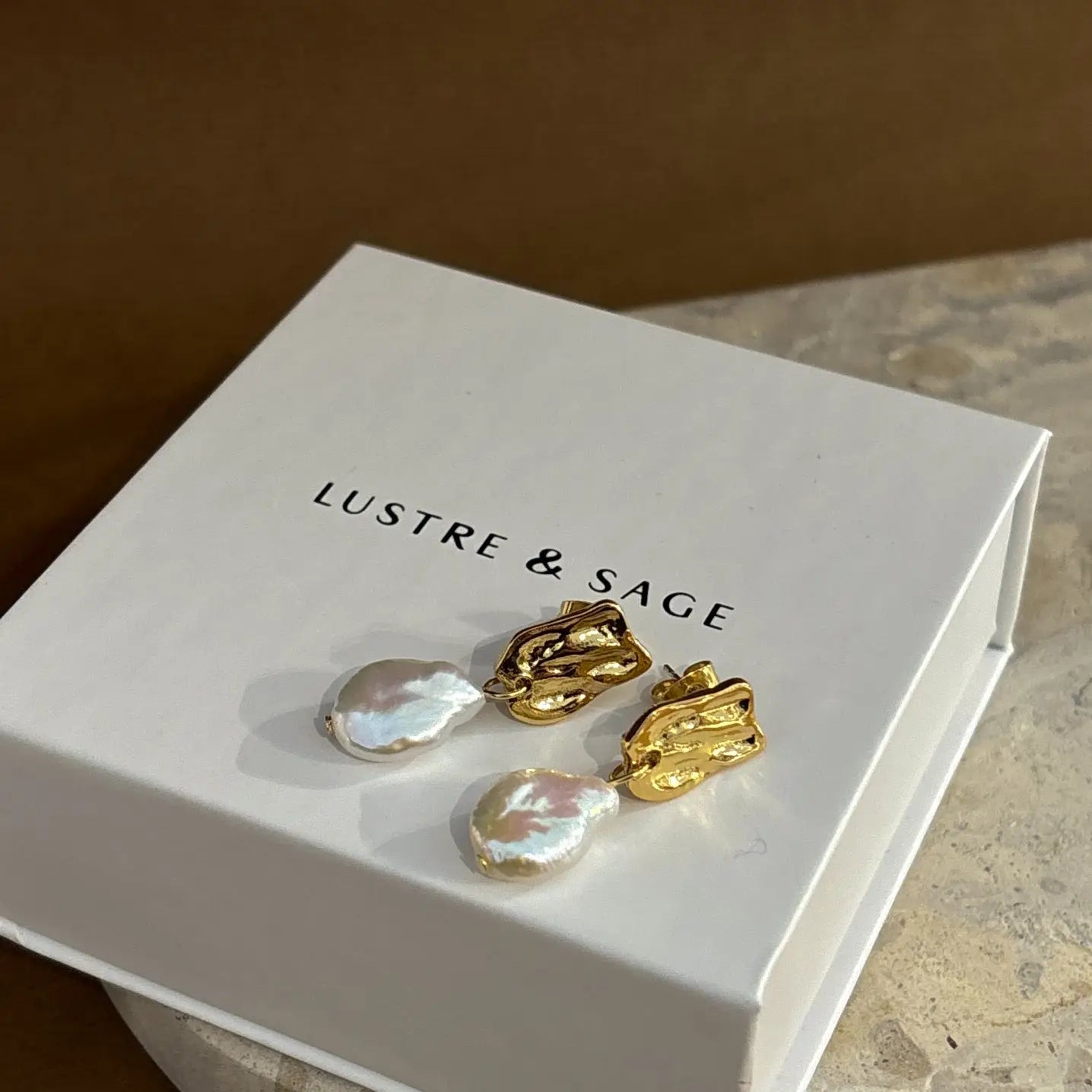 Gold-plated pearl stud earrings displayed on a textured stone surface, highlighting the natural lustre of the pearls.