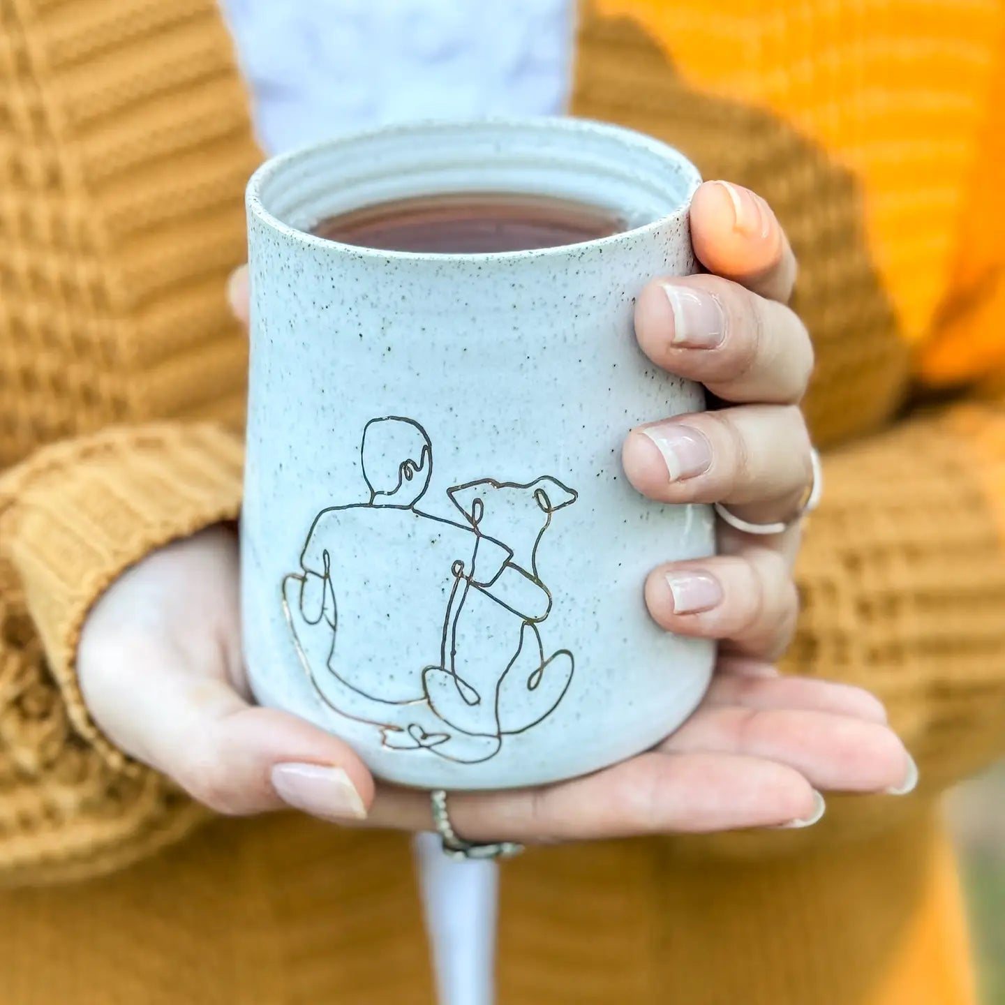 “Person holding a speckled ceramic mug featuring a minimalist line illustration of a man and a dog.”