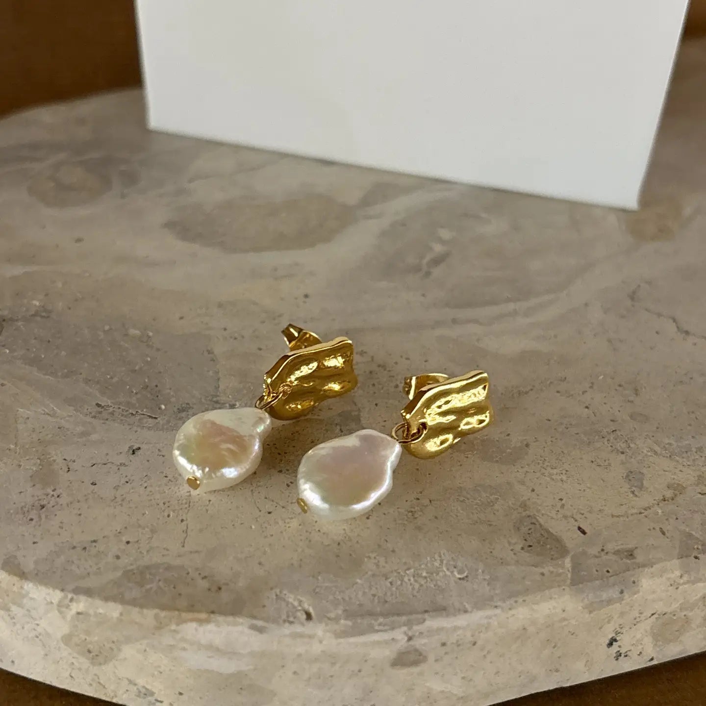 Elegant gold and pearl stud earrings arranged flat on a stone surface, highlighting the organic shape of the pearls.