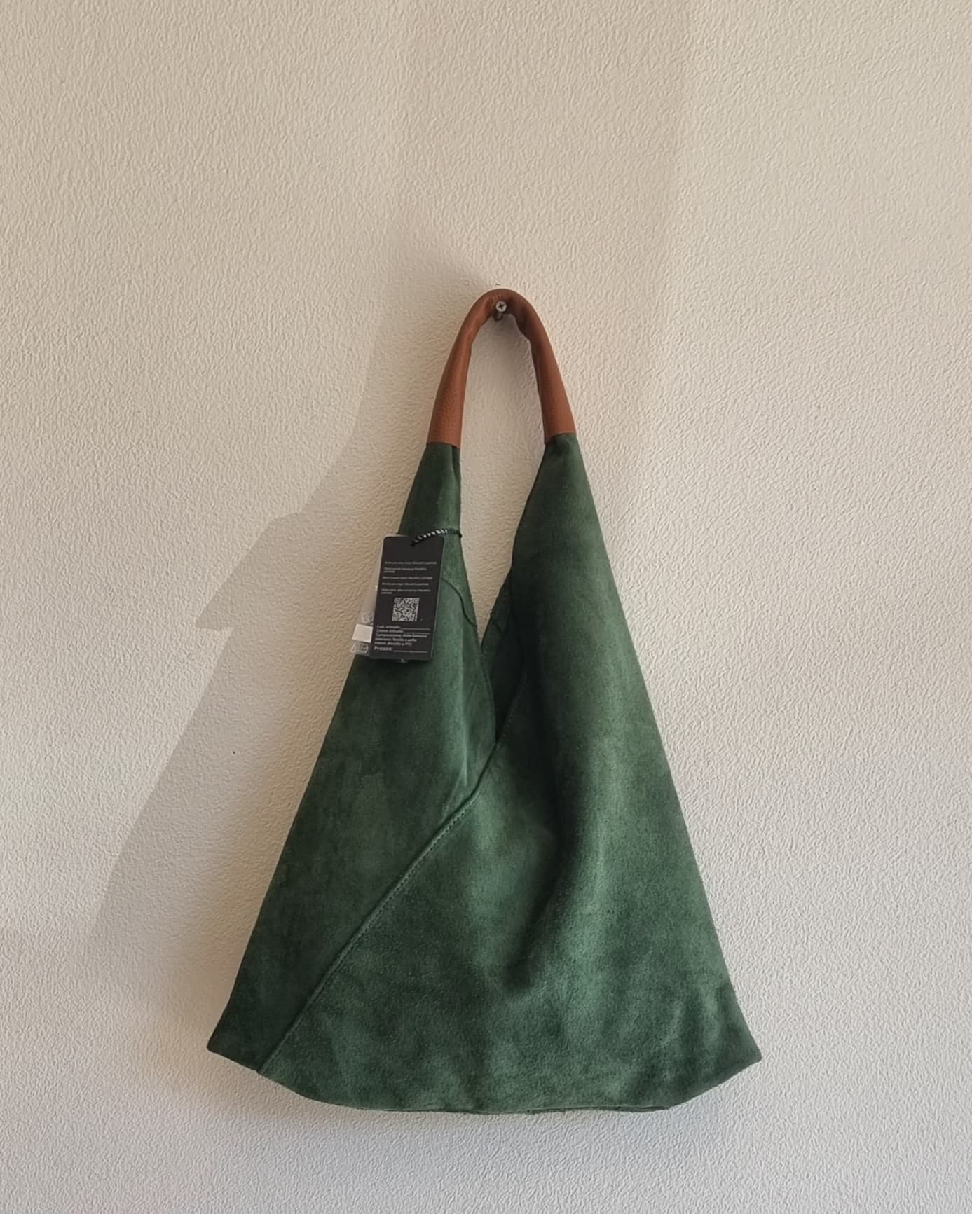 “Green Italian suede shoulder bag with brown handle hanging against a beige textured wall.”