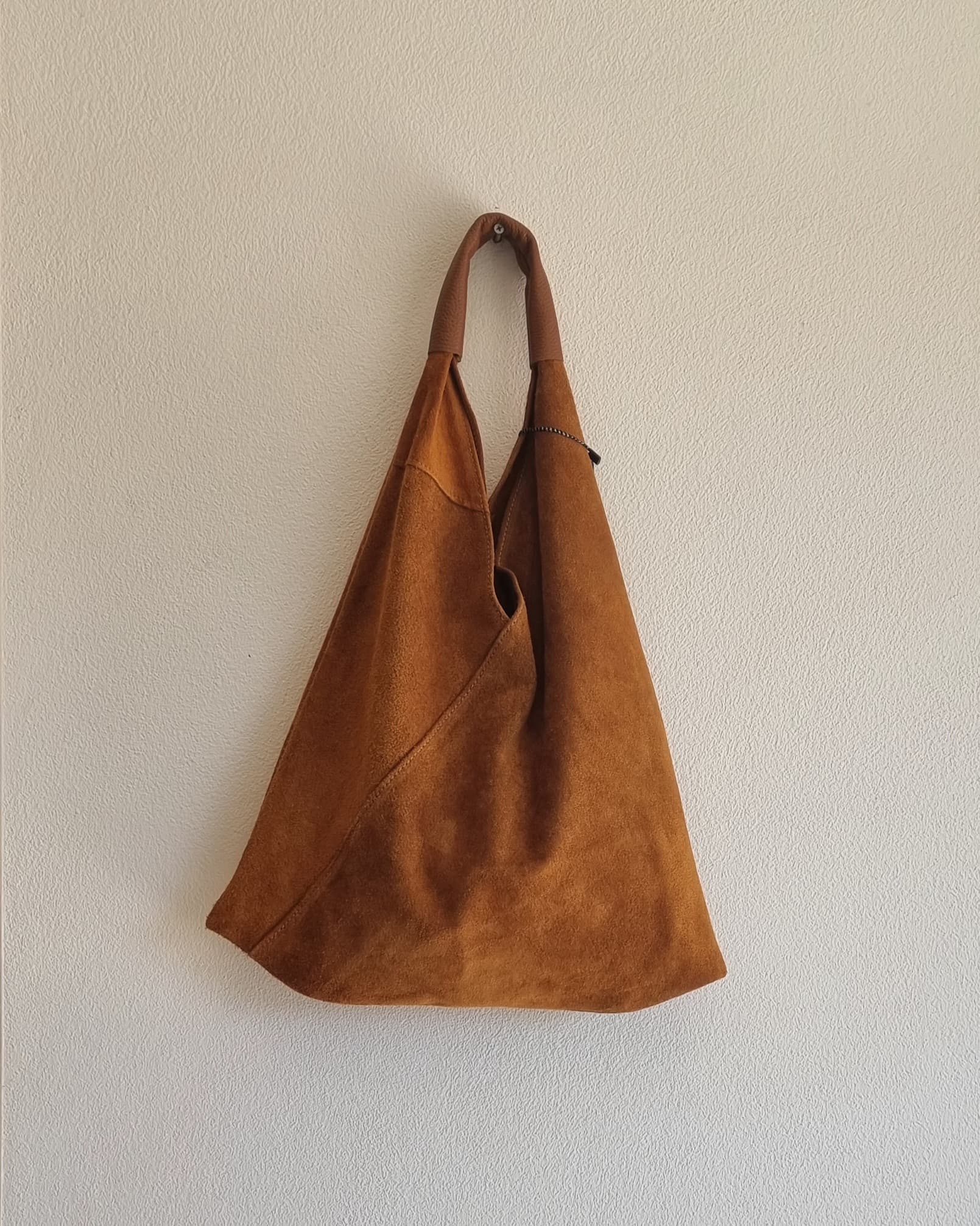 “Brown Italian suede shoulder bag with a brown handle hanging against a beige textured wall.”