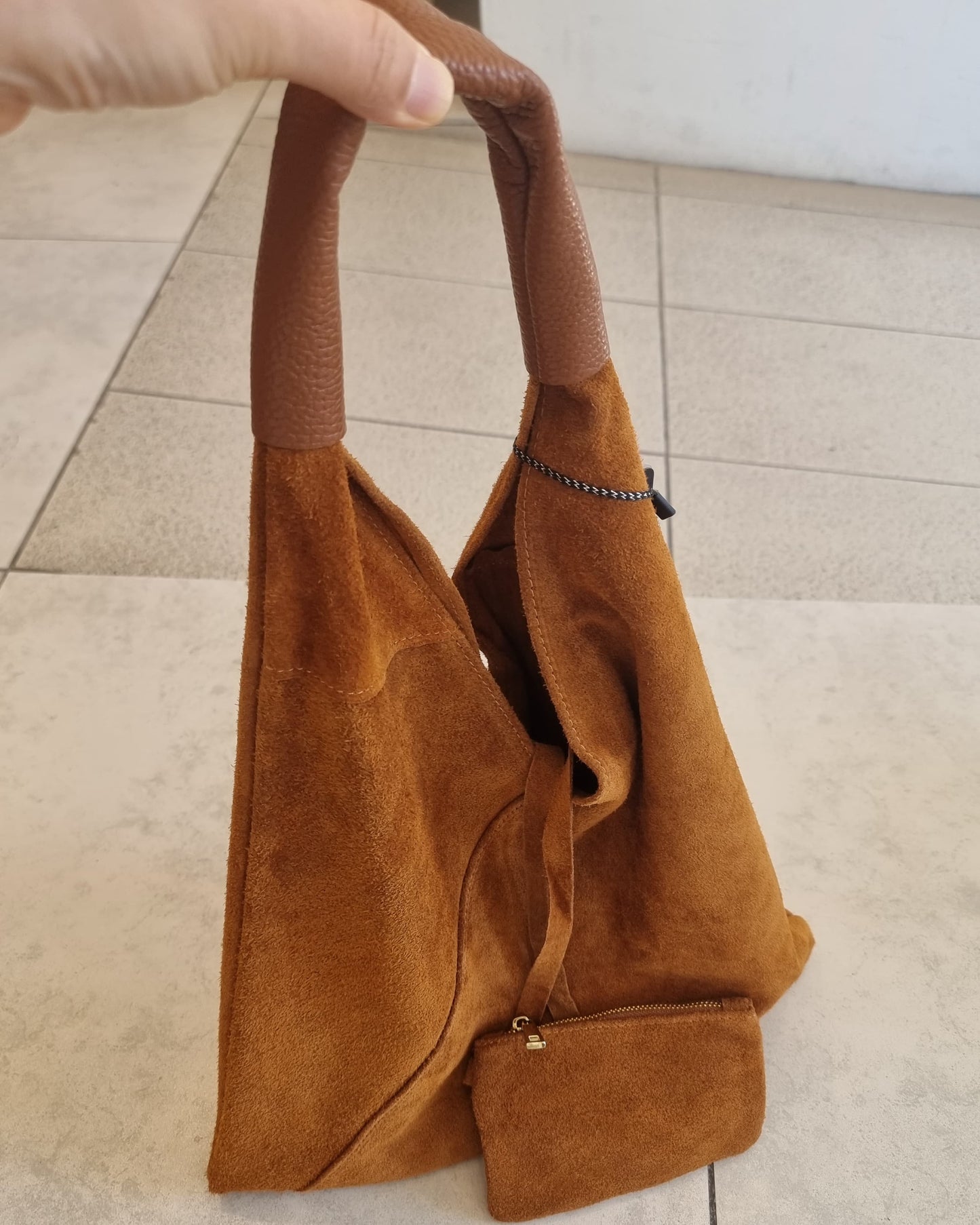 “Brown Italian suede shoulder bag held by hand, featuring a matching zip pouch, shown on a light tiled floor.”