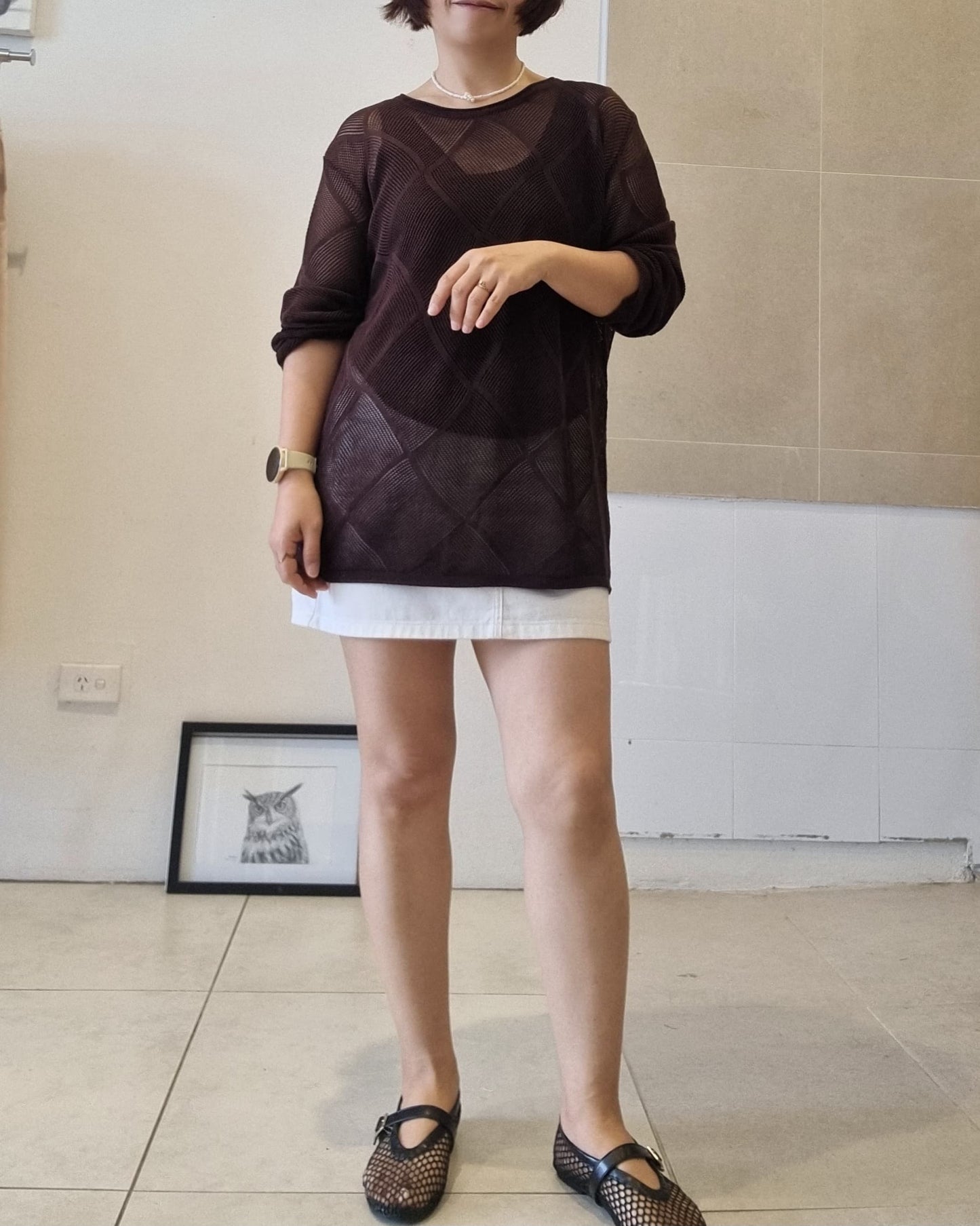 Dark brown sheer knit long-sleeve top styled over a white skirt in a minimal indoor setting