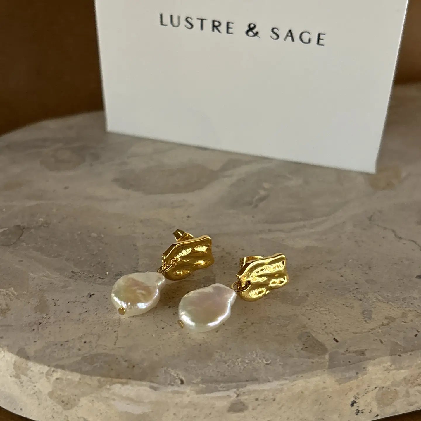 Baroque pearl earrings with gold textured studs presented on a beige marble-like background for product detail.