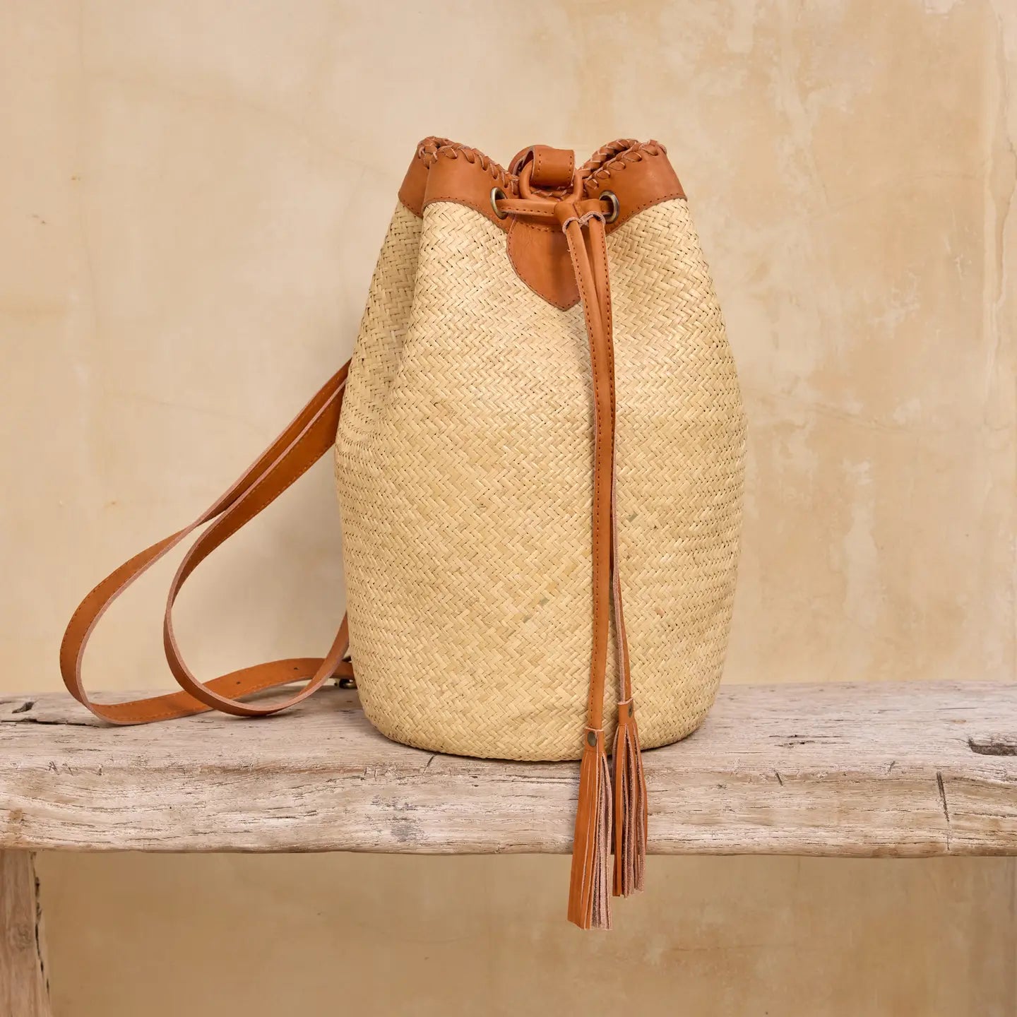 “Handwoven rattan bucket bag with tan leather straps and tassel detail, displayed on a rustic wooden surface.”