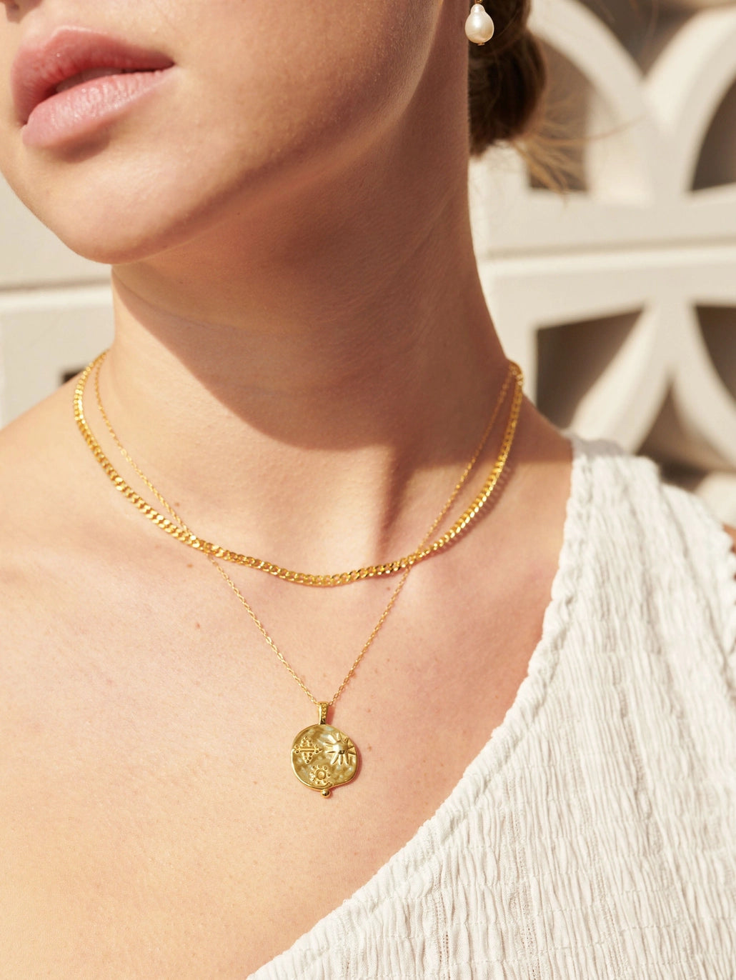Layered gold necklaces including a sun-and-floral medallion pendant styled on a model’s neckline in warm outdoor sunlight.