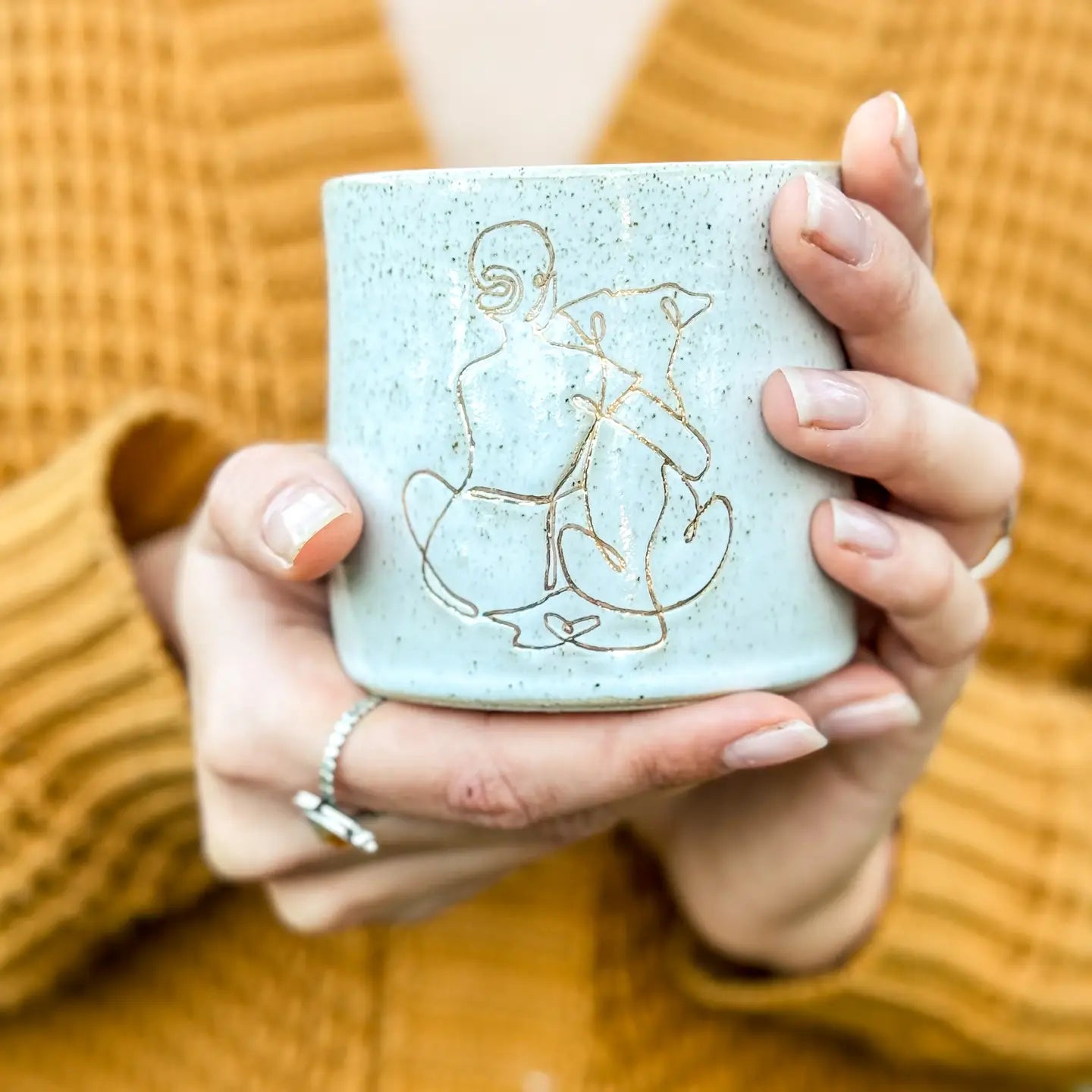 Hands holding a speckled light-blue ceramic mug with a minimalist girl-and-dog gold illustration, against a mustard knit background