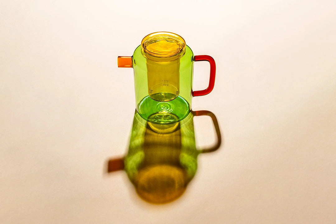 Green glass teapot with yellow lid and orange handle casting a shadow on a beige background