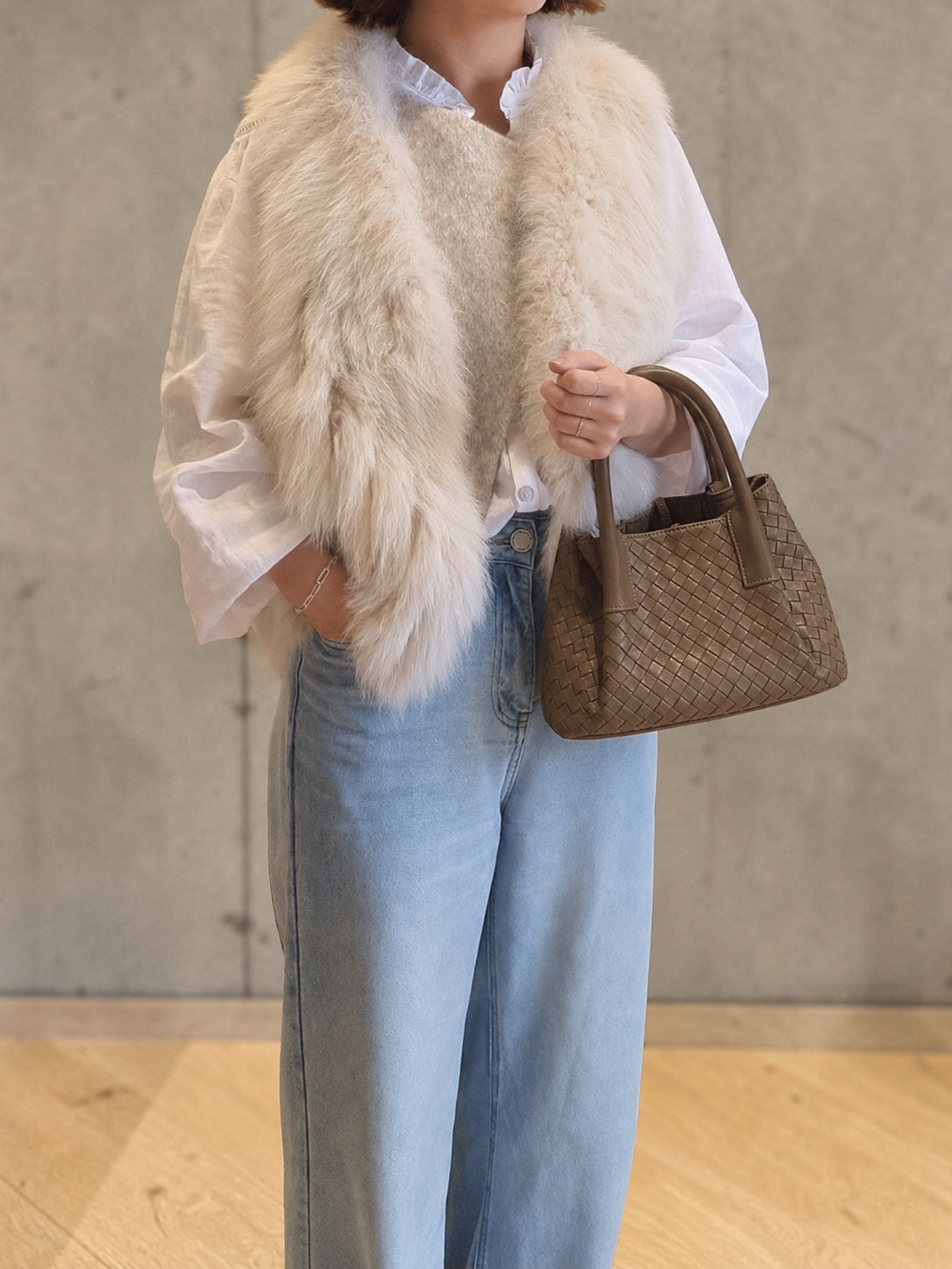 Woman carrying brown woven leather tote bag while wearing cream faux fur vest and light blue jeans