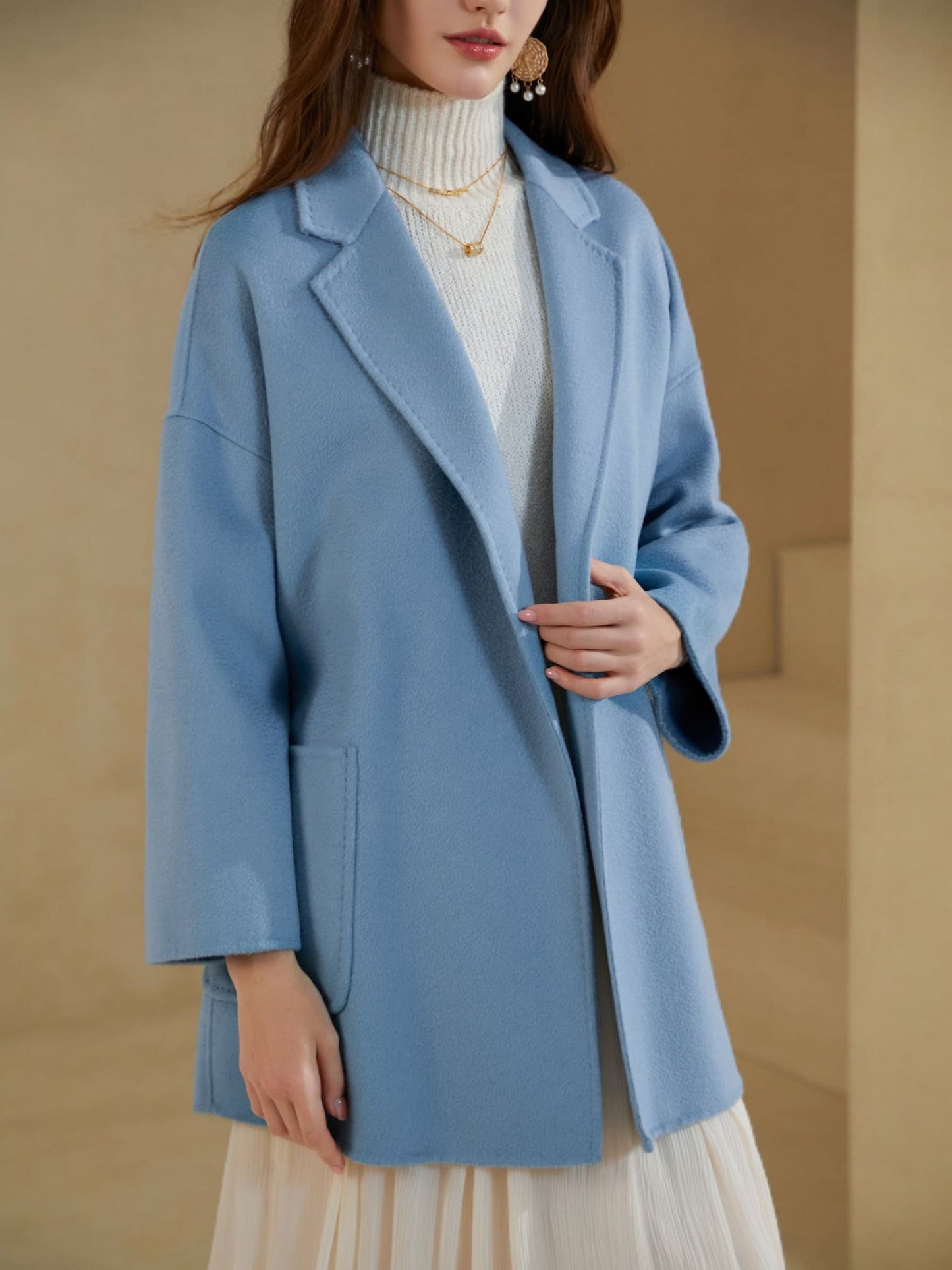 “Close-up of a light blue tailored blazer layered over a white turtleneck knit, paired with an ivory pleated skirt in a minimal neutral setting.”