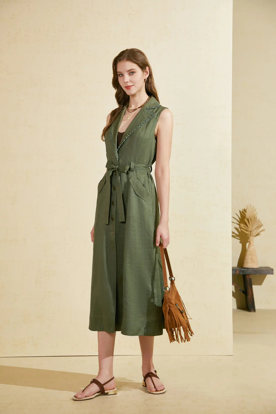Woman wearing a sleeveless green midi dress with a waist tie, holding a brown fringe bag, standing against a beige wall.