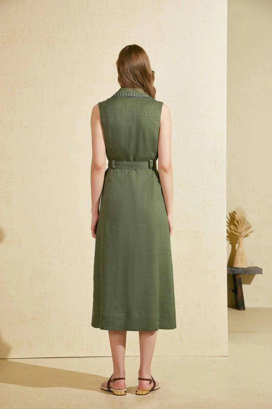 Back view of a sleeveless green long dress with a waist belt, worn by a woman standing against a beige wall.
