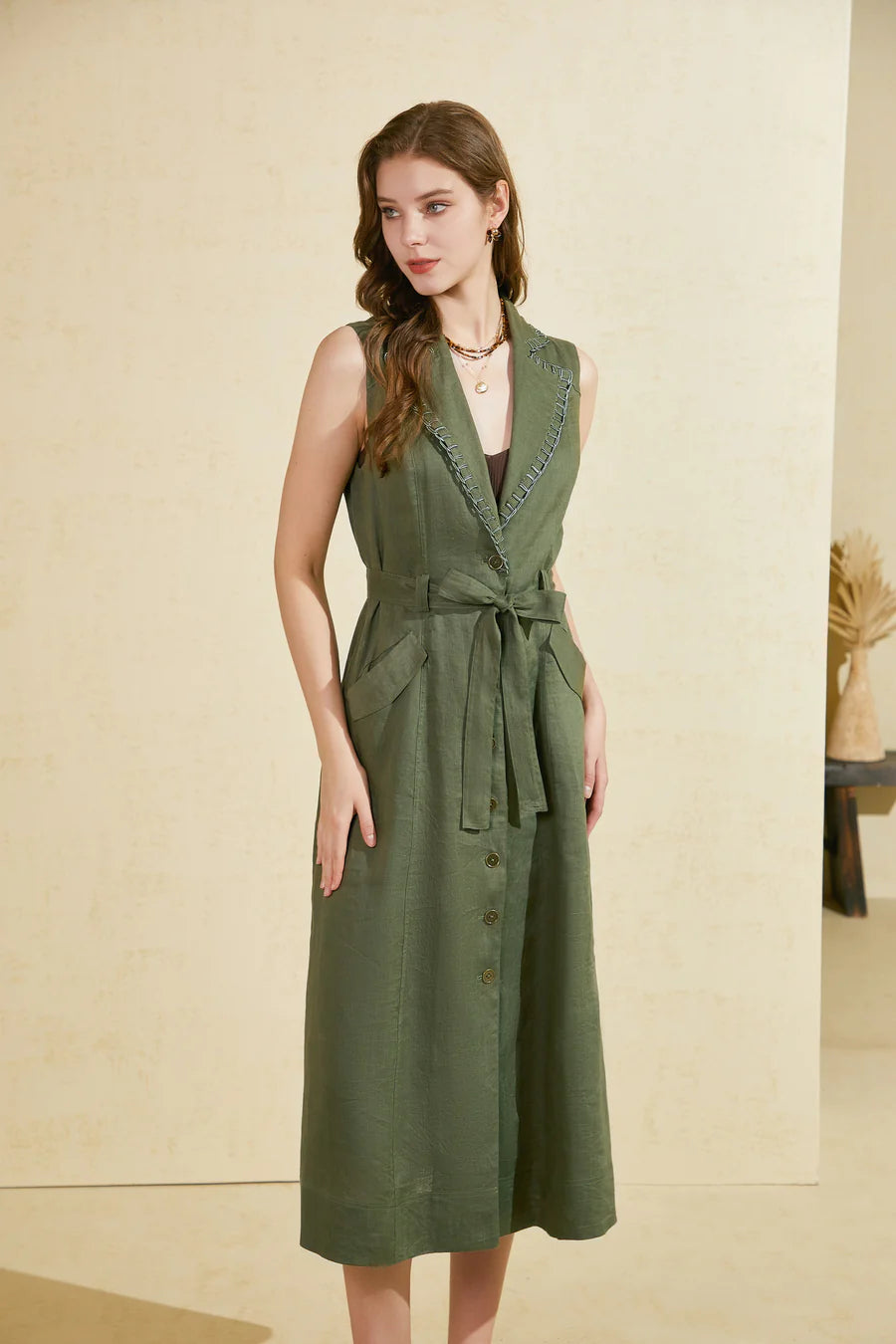 Woman wearing a sleeveless green linen long dress with a waist belt and button-down front, standing against a beige background.
