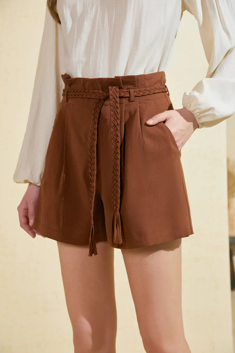 Model wearing brown high-rise belted shorts with pleated front, full-length view