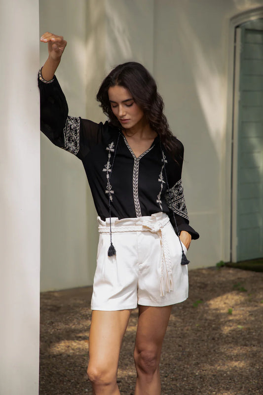 Woman wearing a black embroidered boho blouse with white shorts, leaning lightly against a column