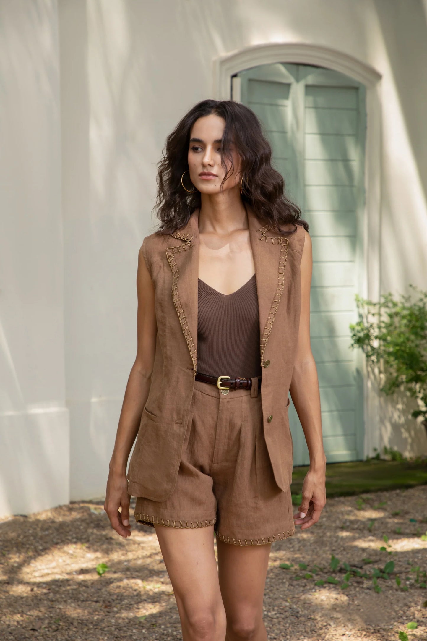 "Woman wearing a brown sleeveless vest and matching shorts walking outdoors in front of a mint green door"
