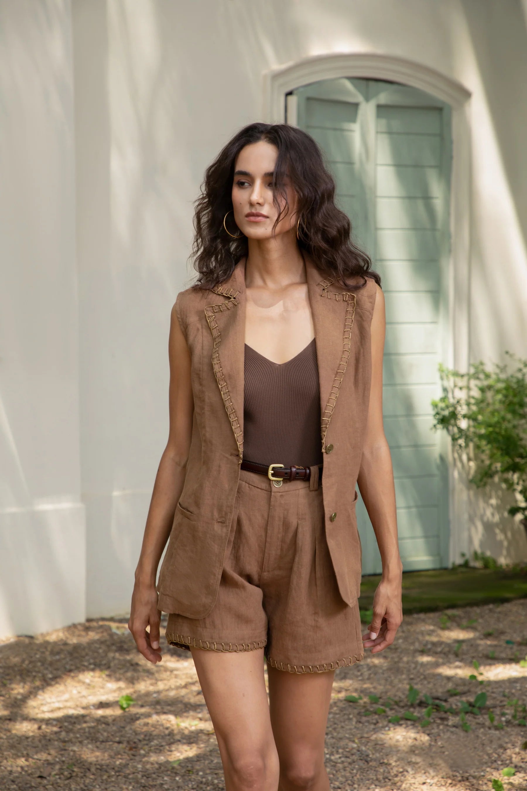 "Woman wearing a brown sleeveless vest and matching shorts walking outdoors in front of a mint green door"