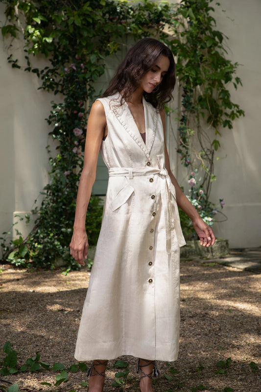 “Woman wearing a beige sleeveless dress with buttons, standing outdoors with greenery in the background.”