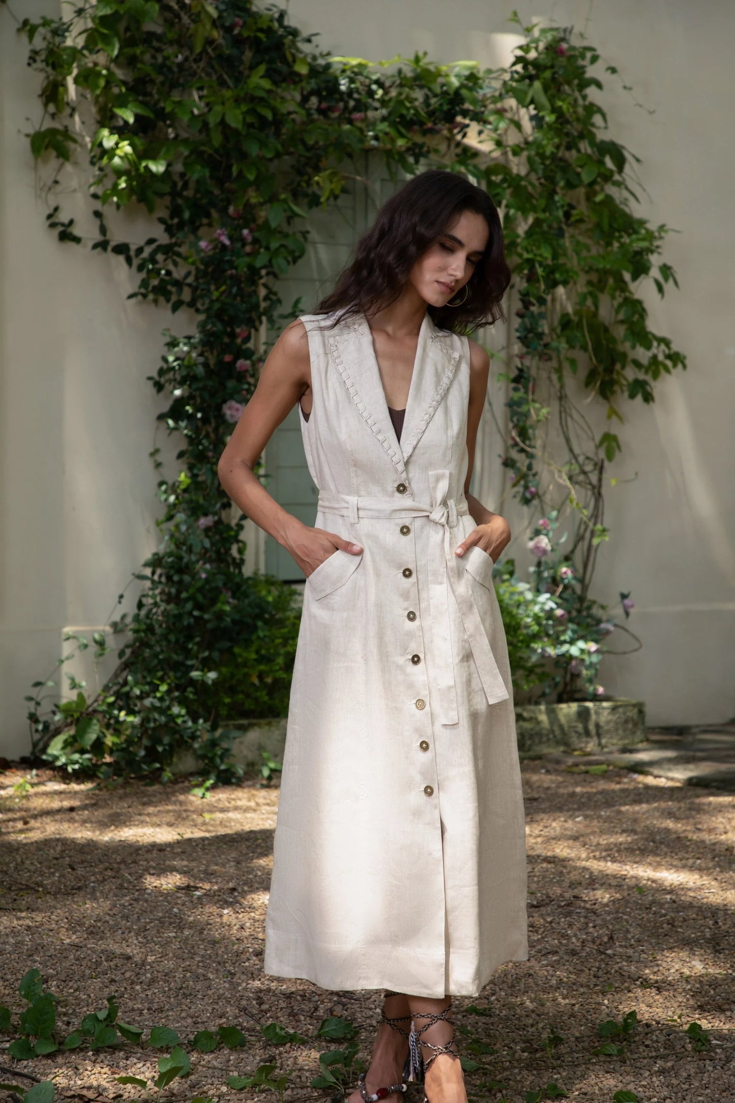 Woman wearing a sleeveless beige button-down midi dress with a waist tie, standing in an outdoor garden with greenery.