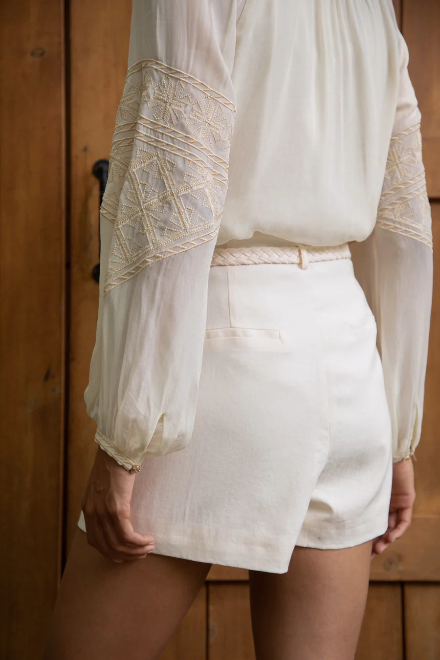 “Close-up view of a woman wearing cream-coloured tailored shorts and an embroidered blouse, standing against a wooden door — Nova Lux.”