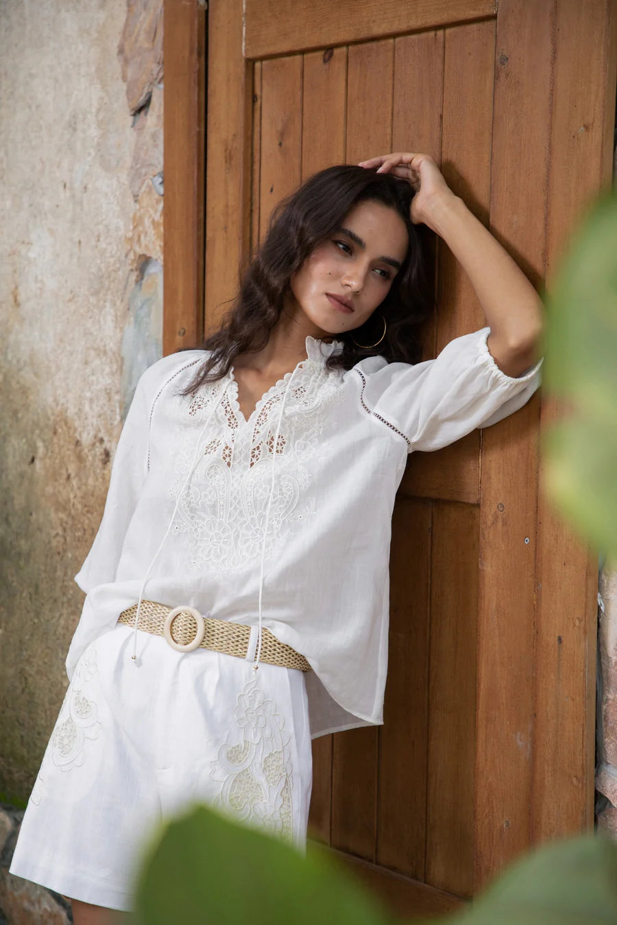 "White embroidered blouse with lace details paired with matching shorts, worn by a woman leaning against a wooden door."