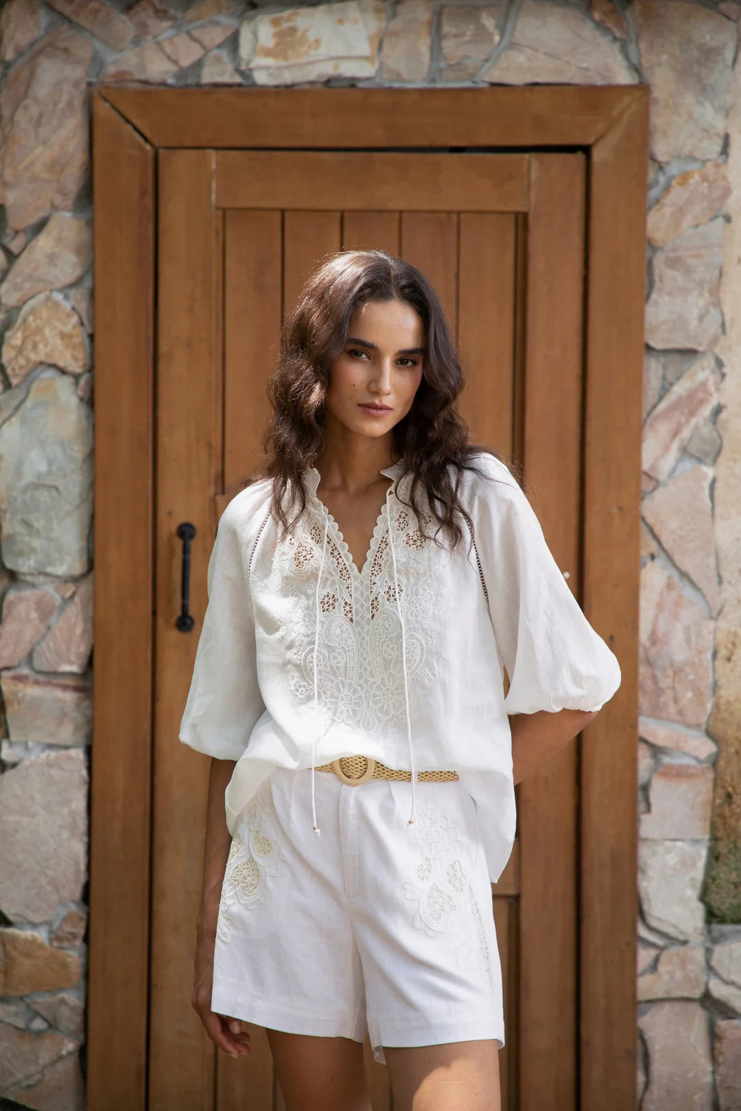 “Model wearing white embroidered high-waist shorts with matching blouse, standing in front of a wooden door.”