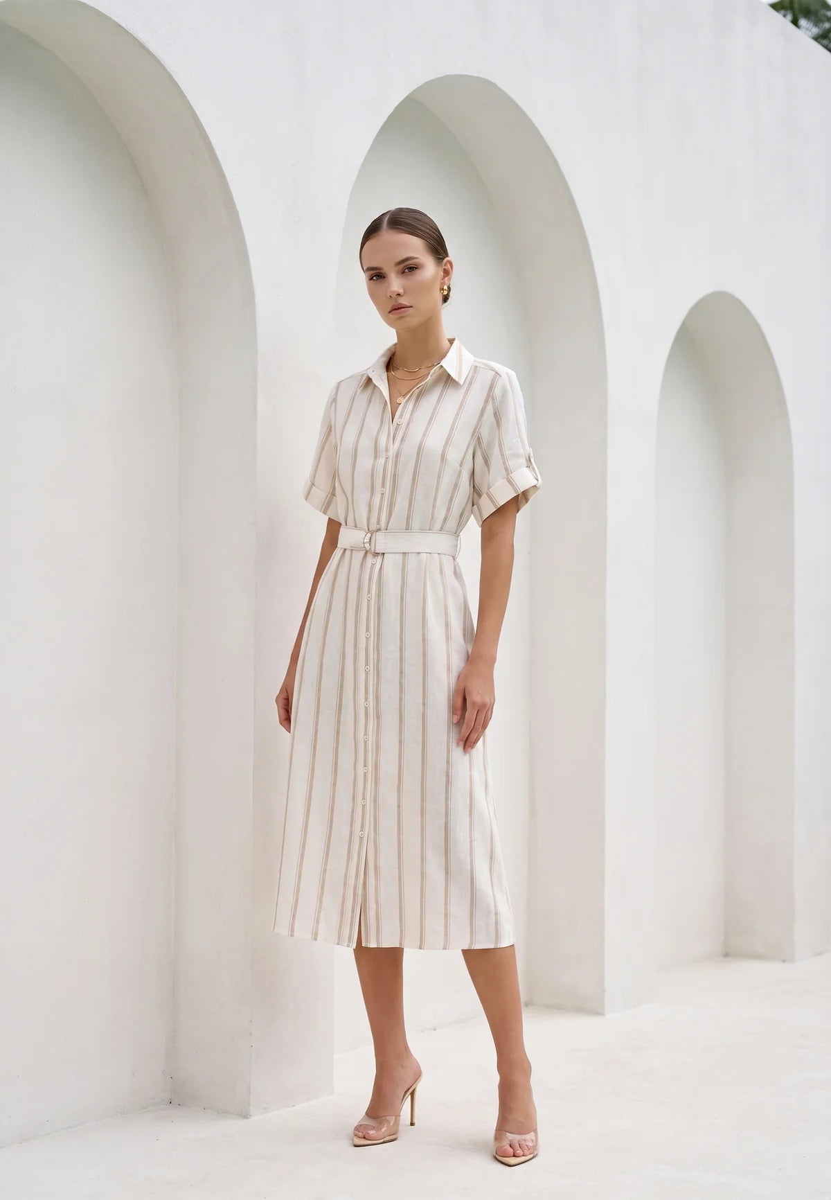 A woman models a short-sleeve collared midi dress with vertical cream and beige stripes, featuring a matching fabric belt at the waist. She is standing in a bright, minimalist white architectural setting with arched alcoves.