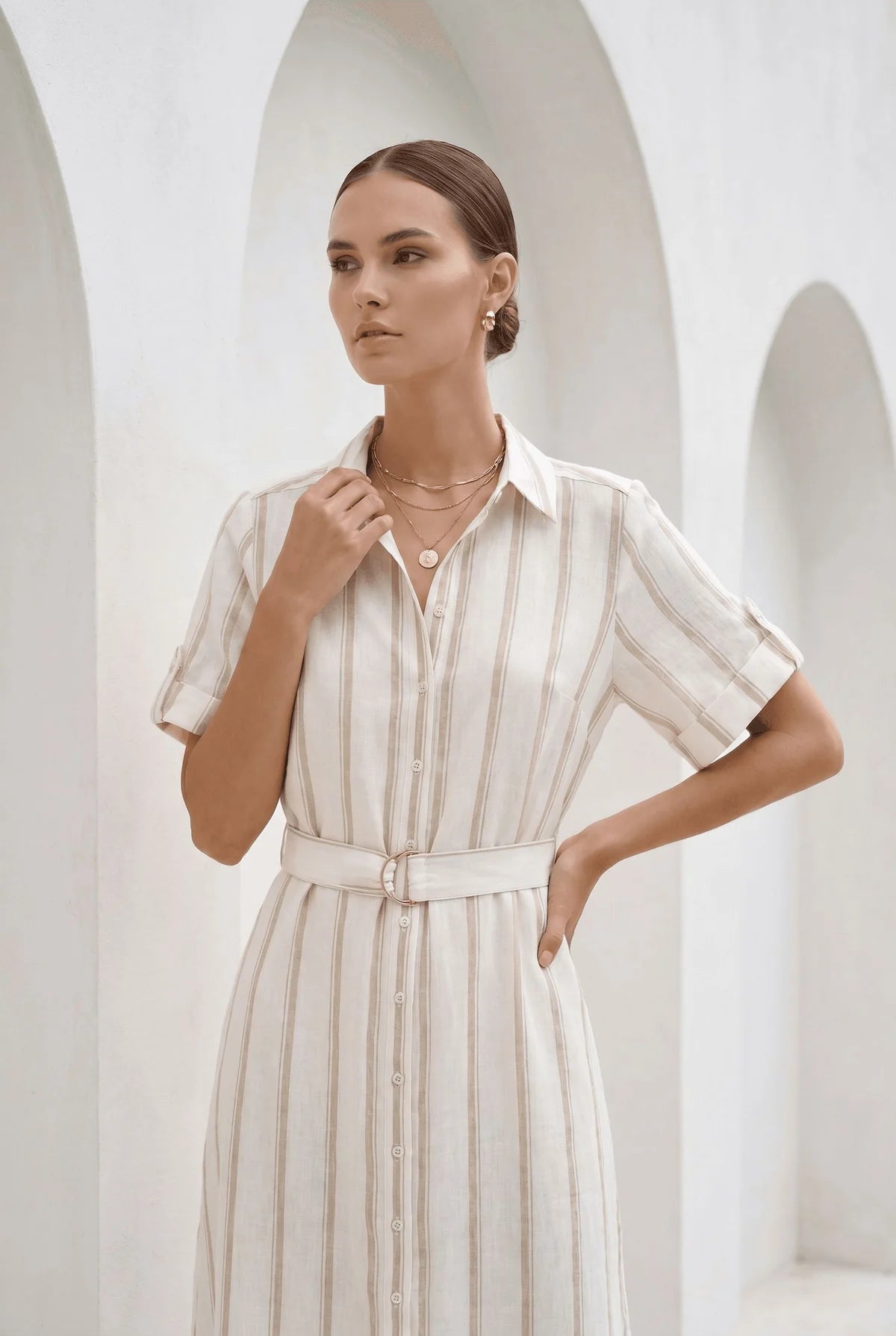 A woman modeling a cream-colored, short-sleeved shirt dress with vertical stripes and a matching belt, set against a white architectural background with arches.