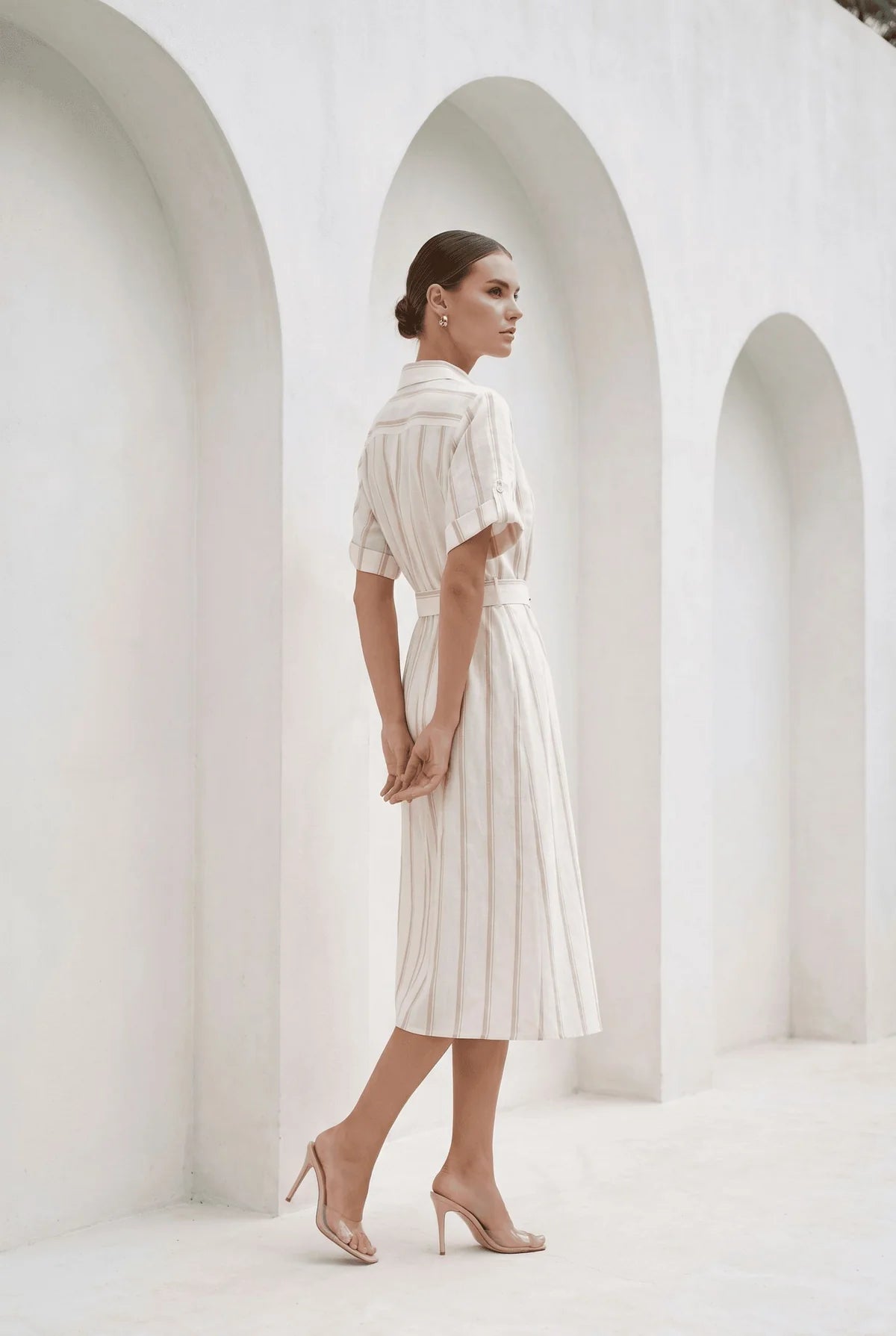A woman stands in profile wearing a cream and white vertically striped short-sleeve midi dress with a tie waist, paired with nude high heels, set against a white architectural background with arches.