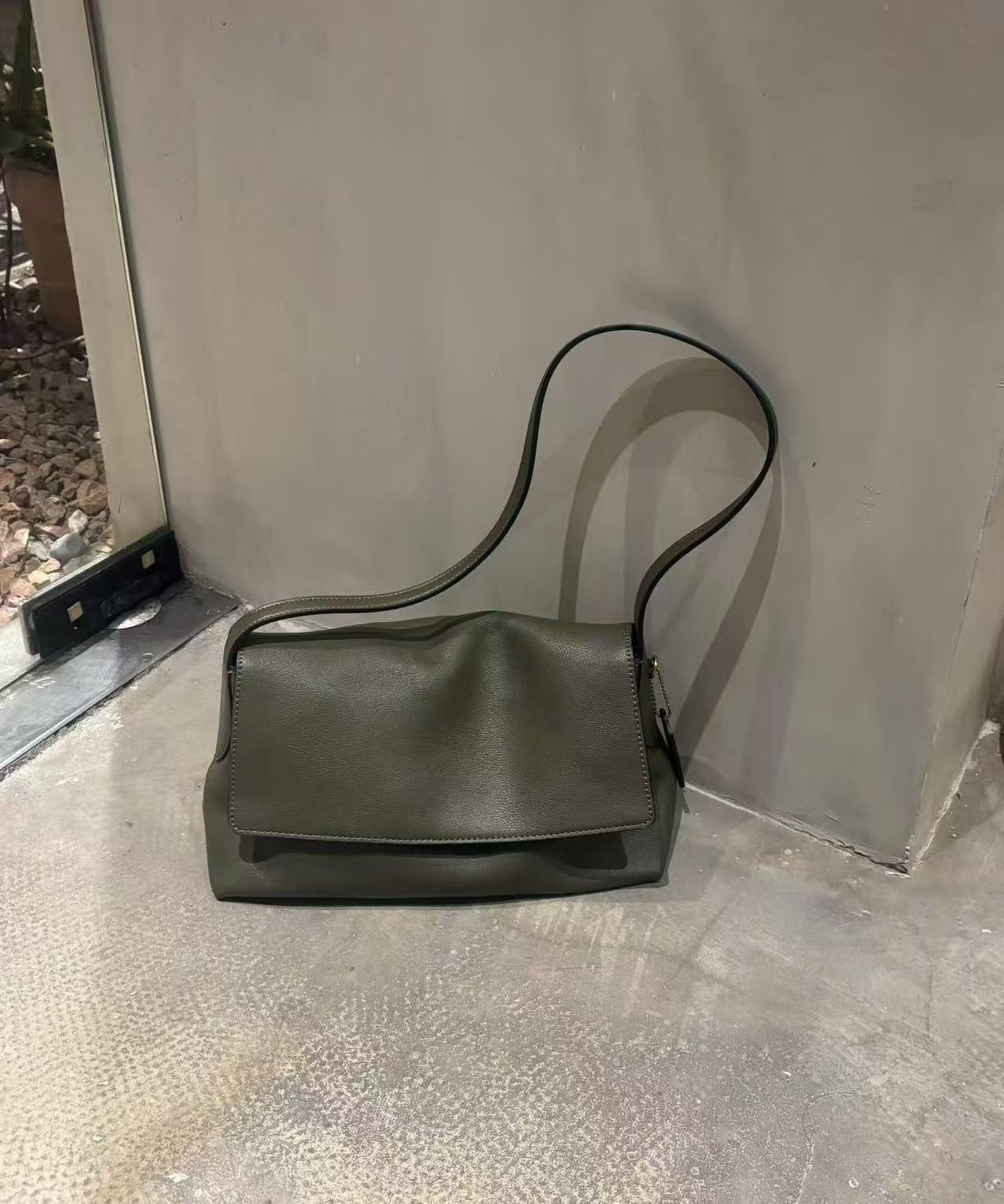 An olive green leather shoulder bag with a long strap, resting on a textured concrete floor against a gray concrete wall.