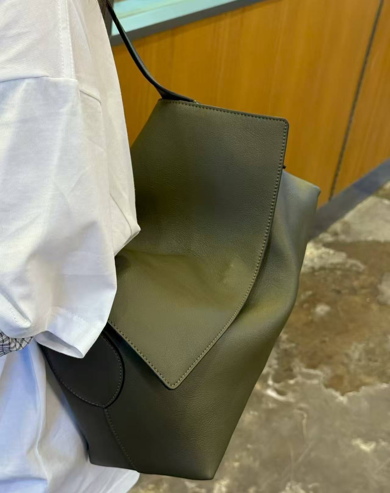 A person wearing a white shirt carries a large olive green leather shoulder bag with a structured flap and black trim, photographed in an indoor setting with a wood-paneled wall and stone floor visible.