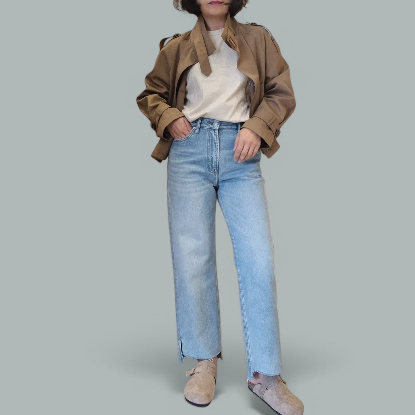 "Woman wearing a beige cropped trench coat over a white top with light blue jeans on a gray background."
