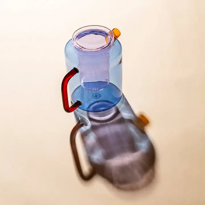 Blue glass Cha Pot with a red-orange handle and yellow spout, photographed on a beige background