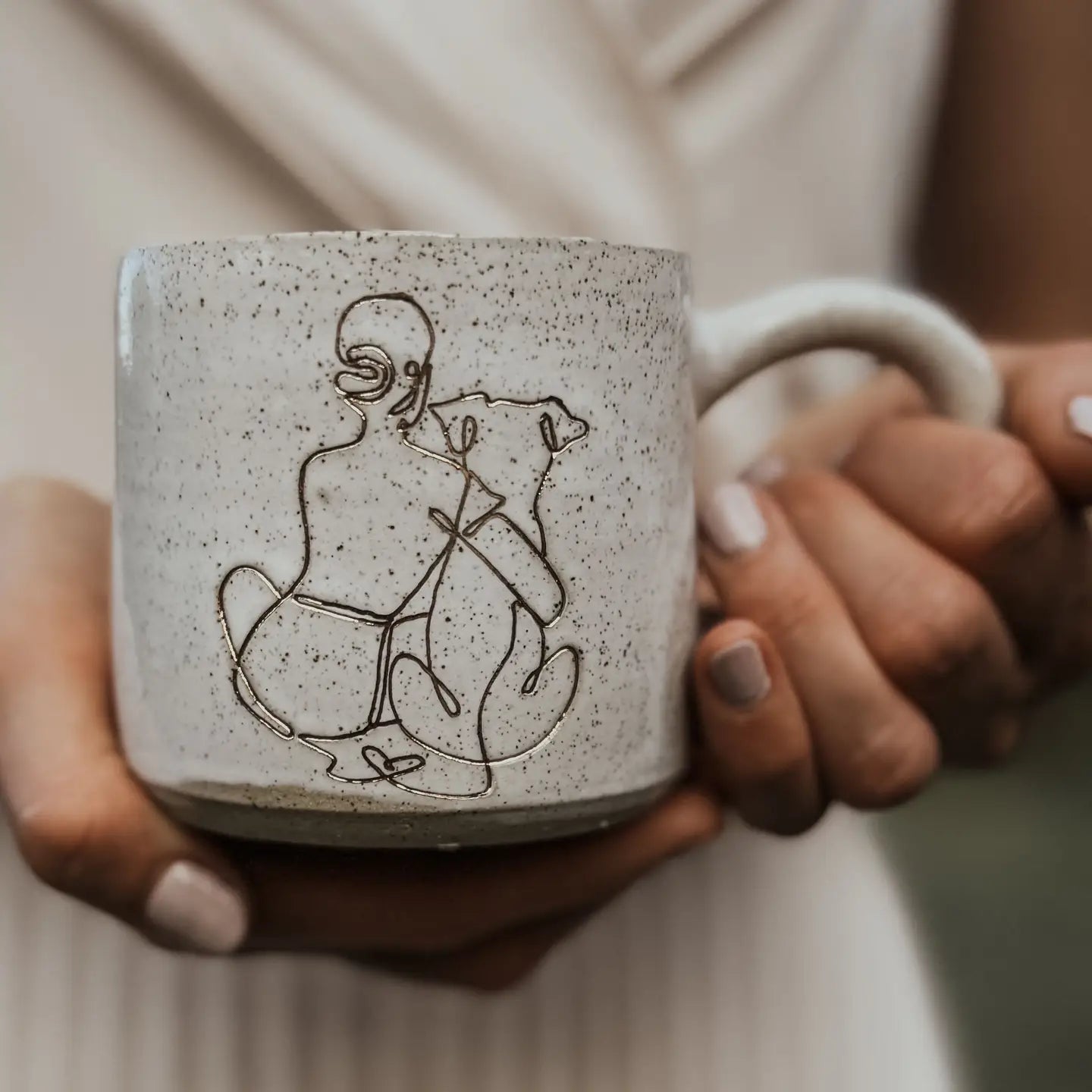 Speckled ceramic mug featuring a minimalist girl-and-dog illustration, held in hands against a soft neutral background