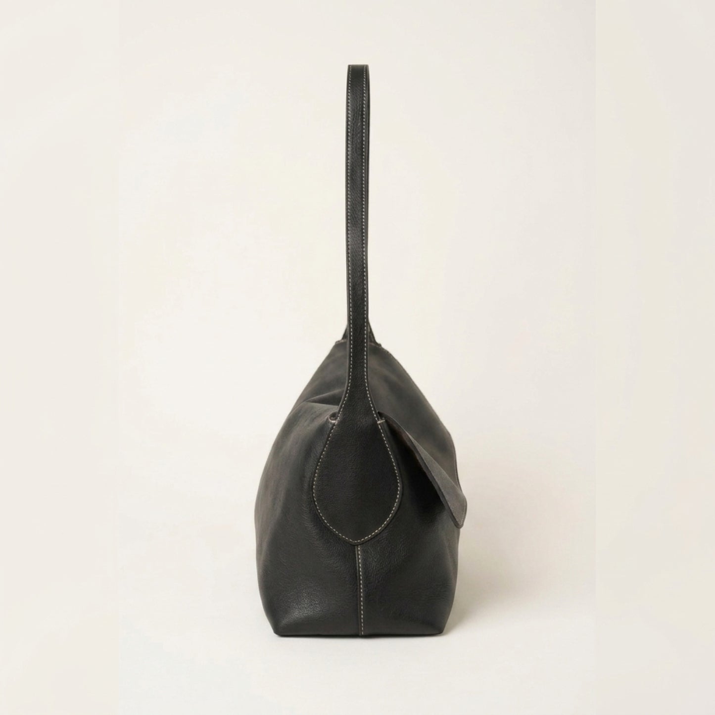 A black leather shoulder bag with a single strap and a small circular leather detail on the front, shown from the side against a plain off-white background.