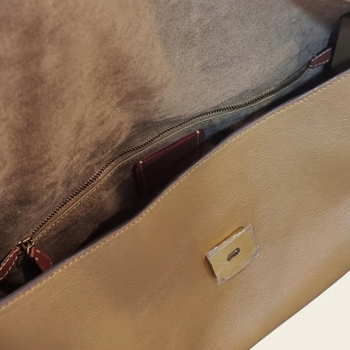Close-up of a tan leather clutch or envelope-style handbag showing the interior zip pocket with a brown suede-like lining and a small gold-tone metal accent on the front.