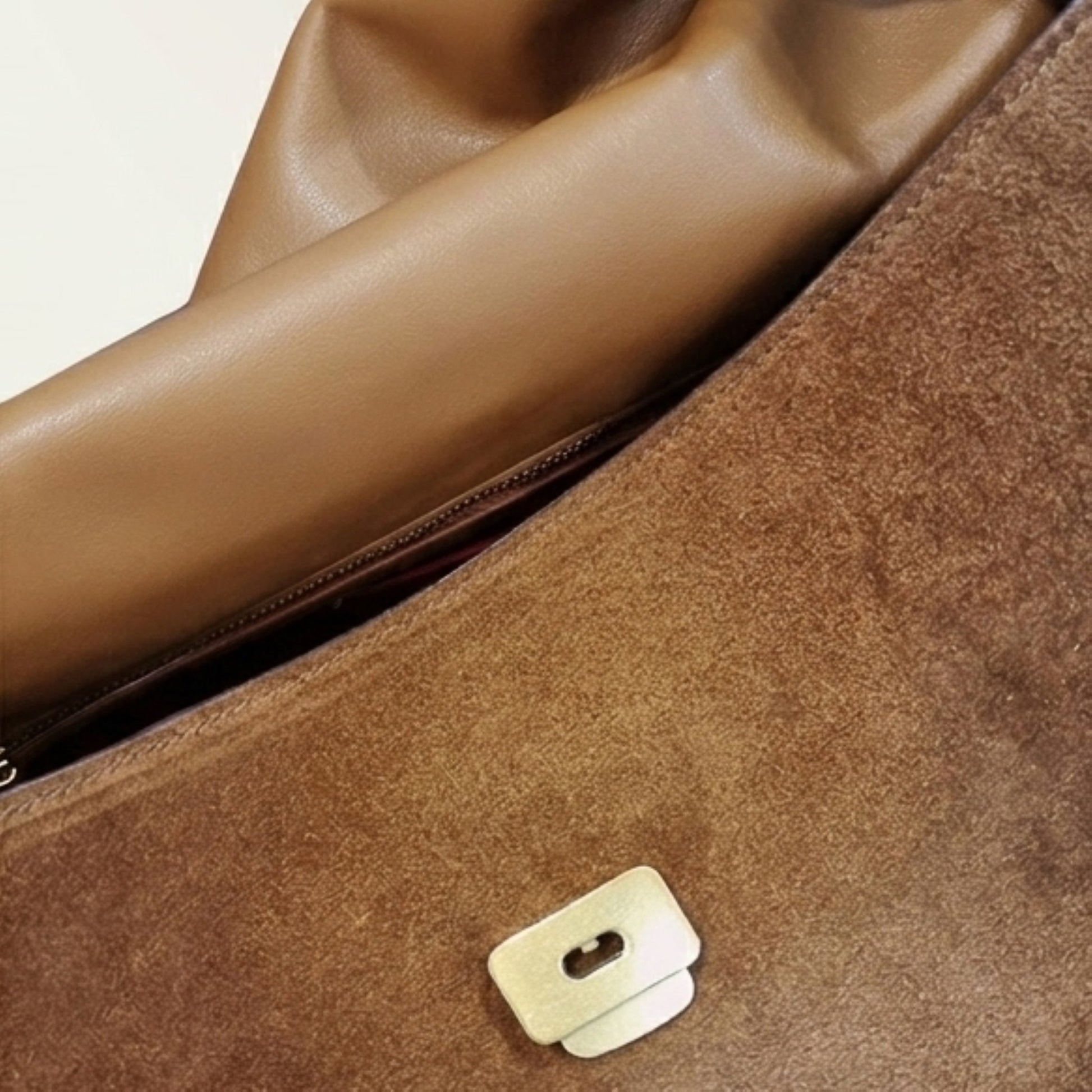 Close-up of a brown suede or faux-suede bag surface with a small silver metal clasp and part of a tan leather zipper interior.