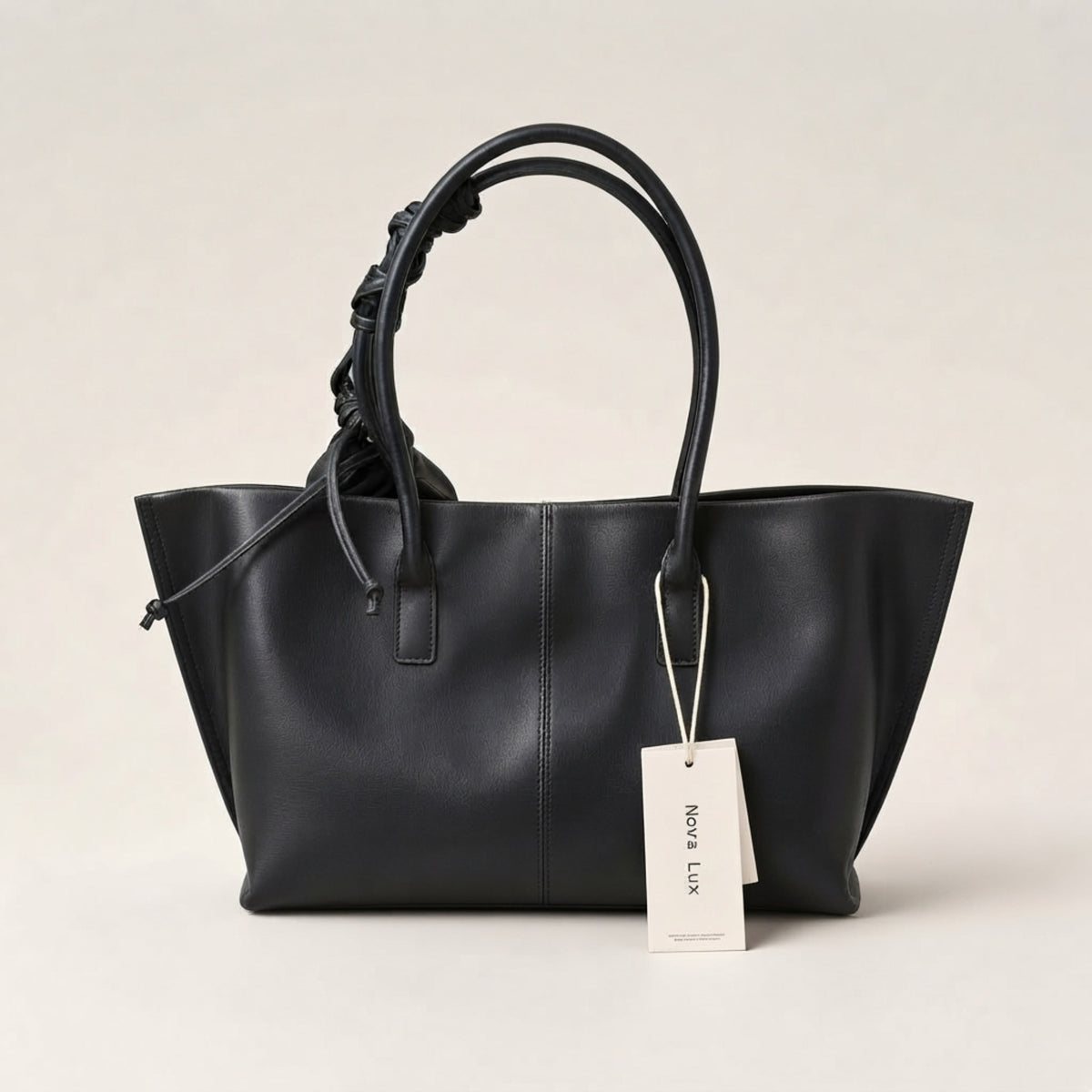 A black leather tote bag with double top handles, one of which is knotted, shown with a white paper tag that reads 'NORA LUX' against a light beige background.