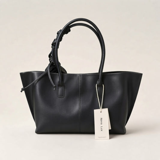 A black leather tote bag with double top handles, one of which is knotted, shown with a white paper tag that reads 'NORA LUX' against a light beige background.