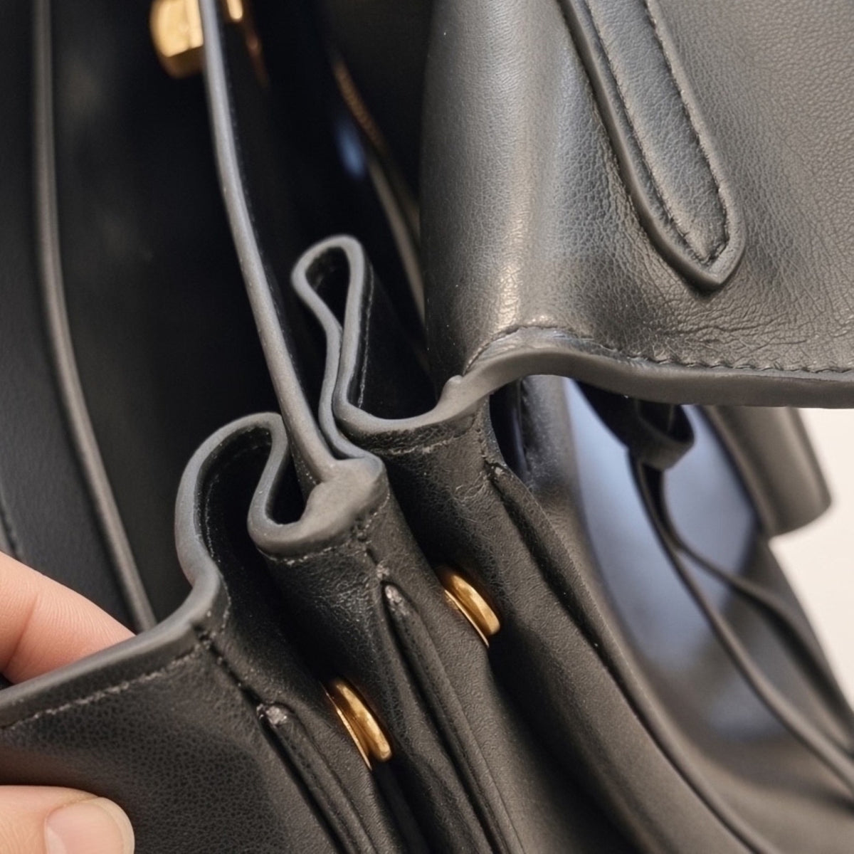Close-up of a black leather handbag with gold-tone hardware and a hand holding the flap open to show the interior lining and drawstring details.