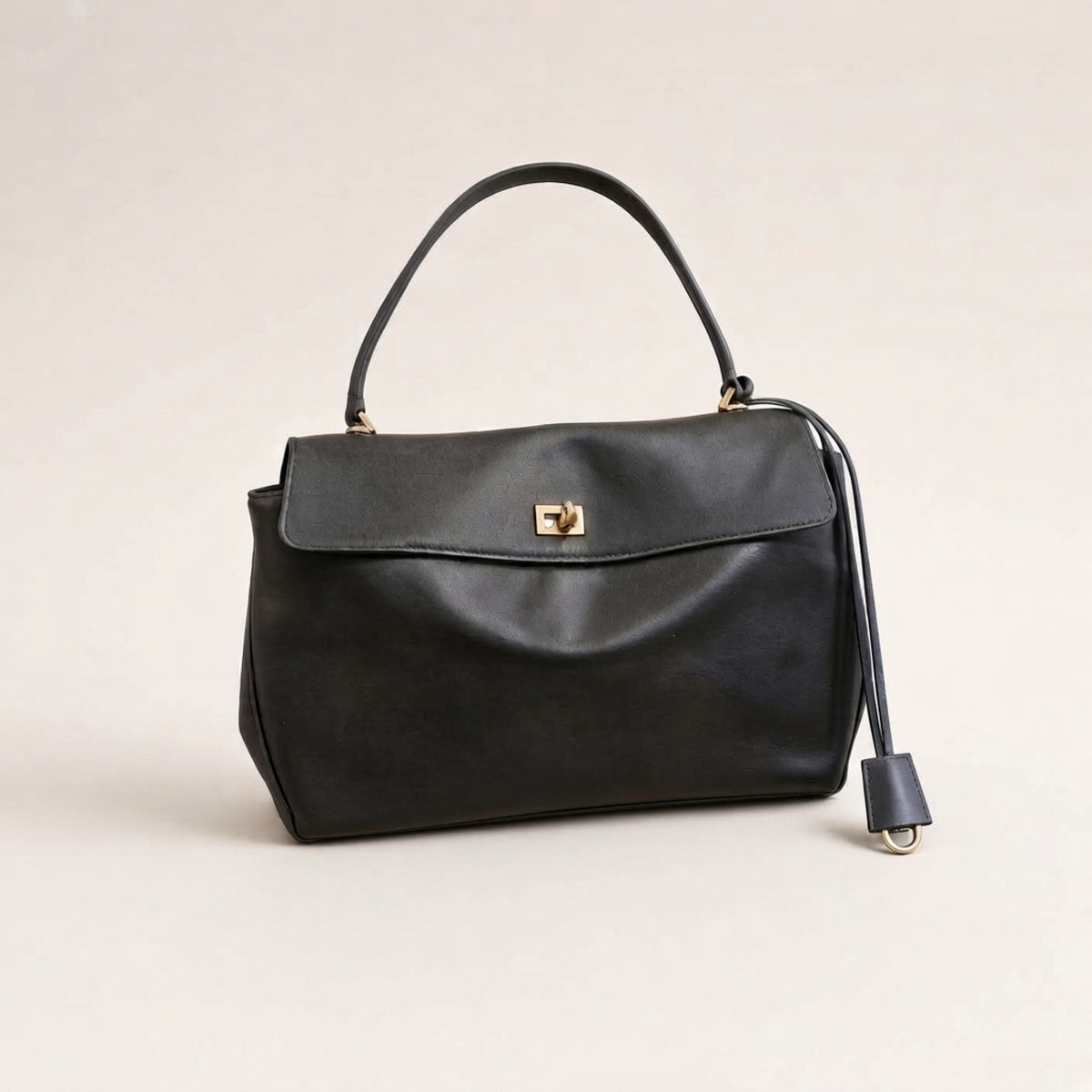 A black leather handbag with a top handle, a front flap featuring a gold-tone turn-lock clasp, and a detachable shoulder strap, photographed on a neutral light beige background.