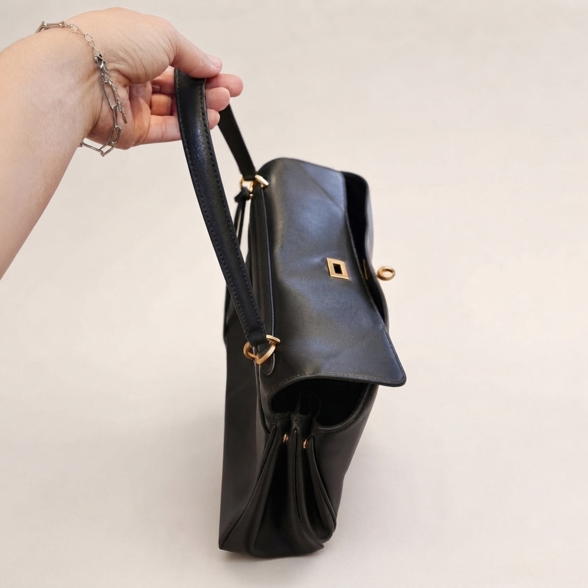 A black leather handbag with gold-tone hardware, held by a person's hand against a plain light beige background.