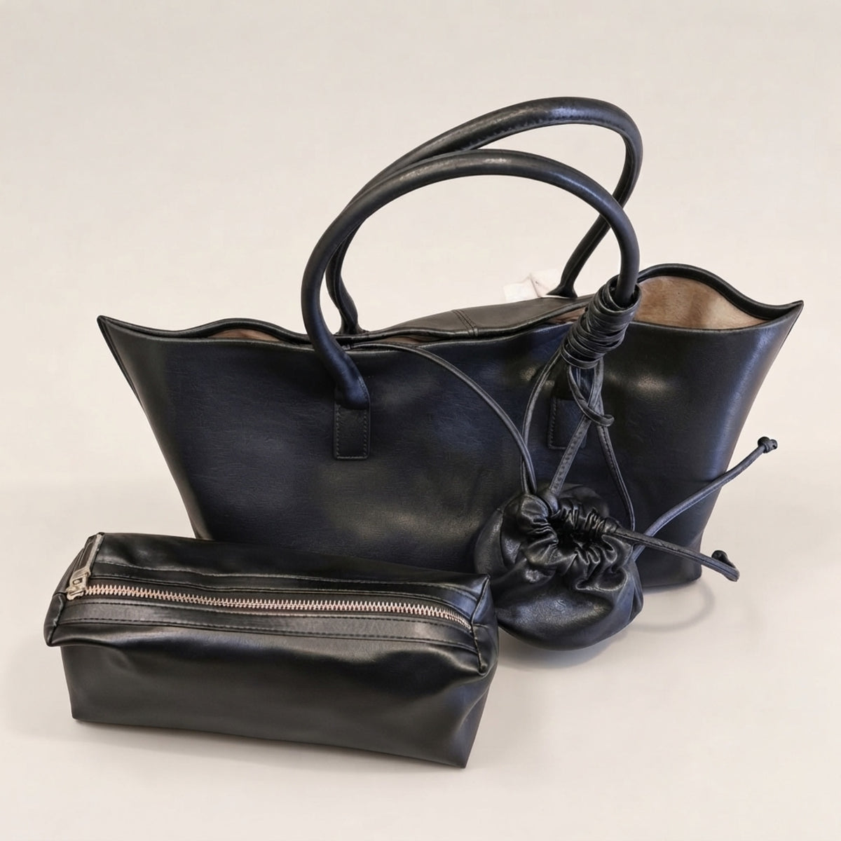 A black leather tote bag with dual top handles, a matching zippered pouch, and a small black drawstring pouch, displayed on a light gray background.