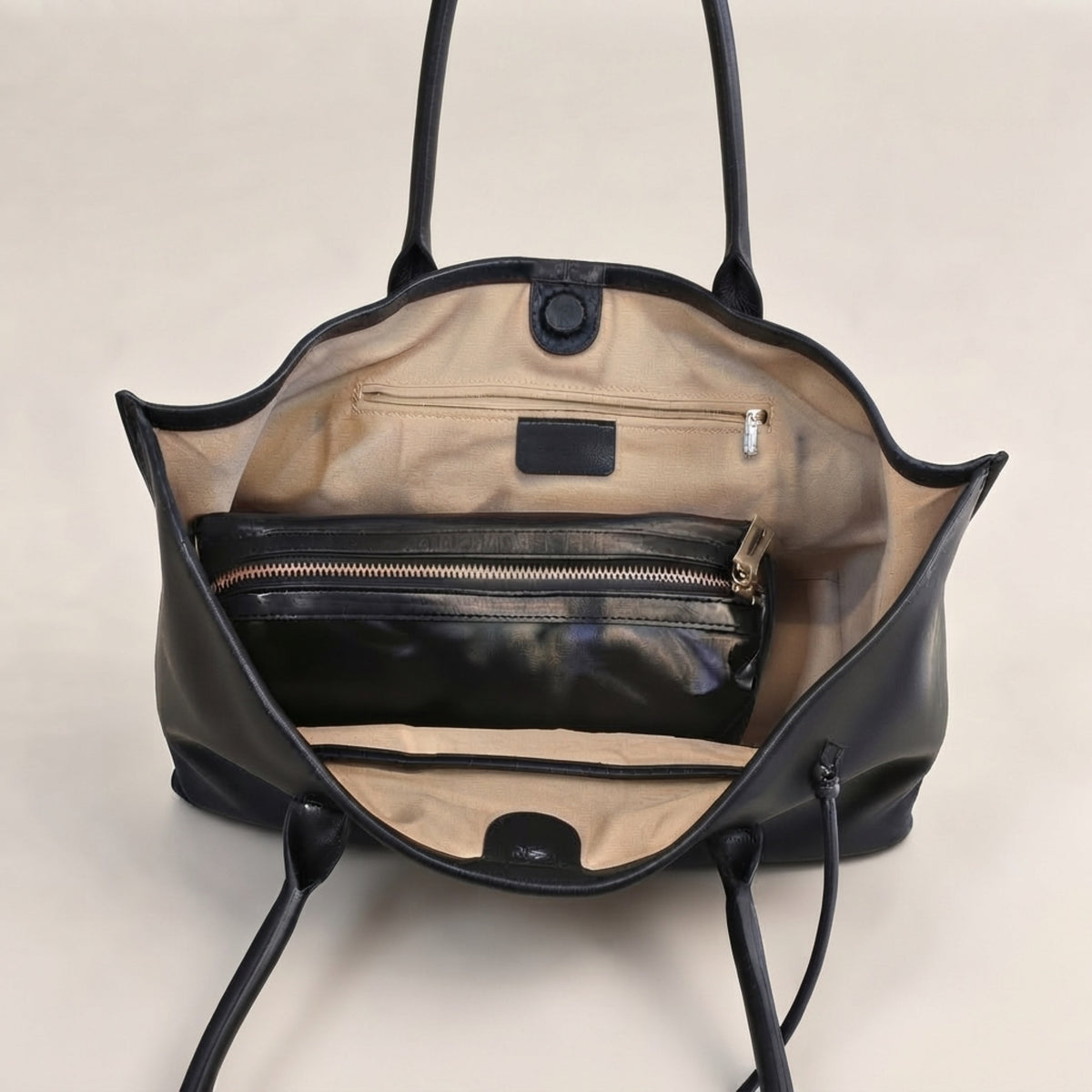 An open black leather tote bag showing its beige interior lining, including a zippered pocket, a coin pocket, and a small black brand plaque.