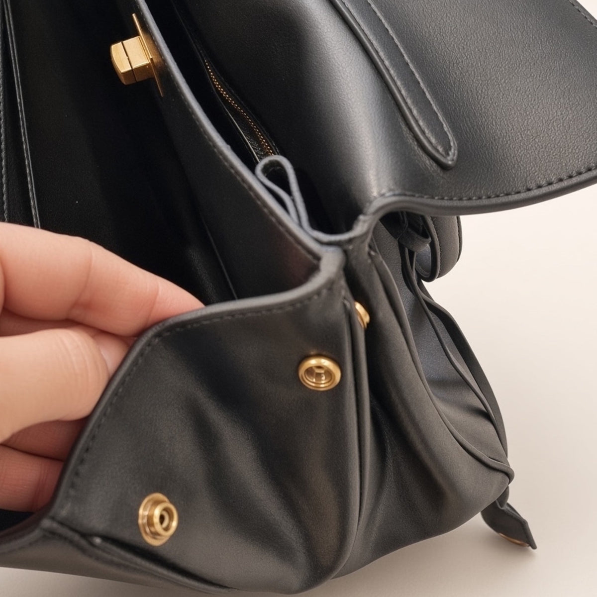 Close-up of a black leather handbag with gold-tone hardware, showing a hand holding open one of the front flap pockets to reveal the interior zipper.