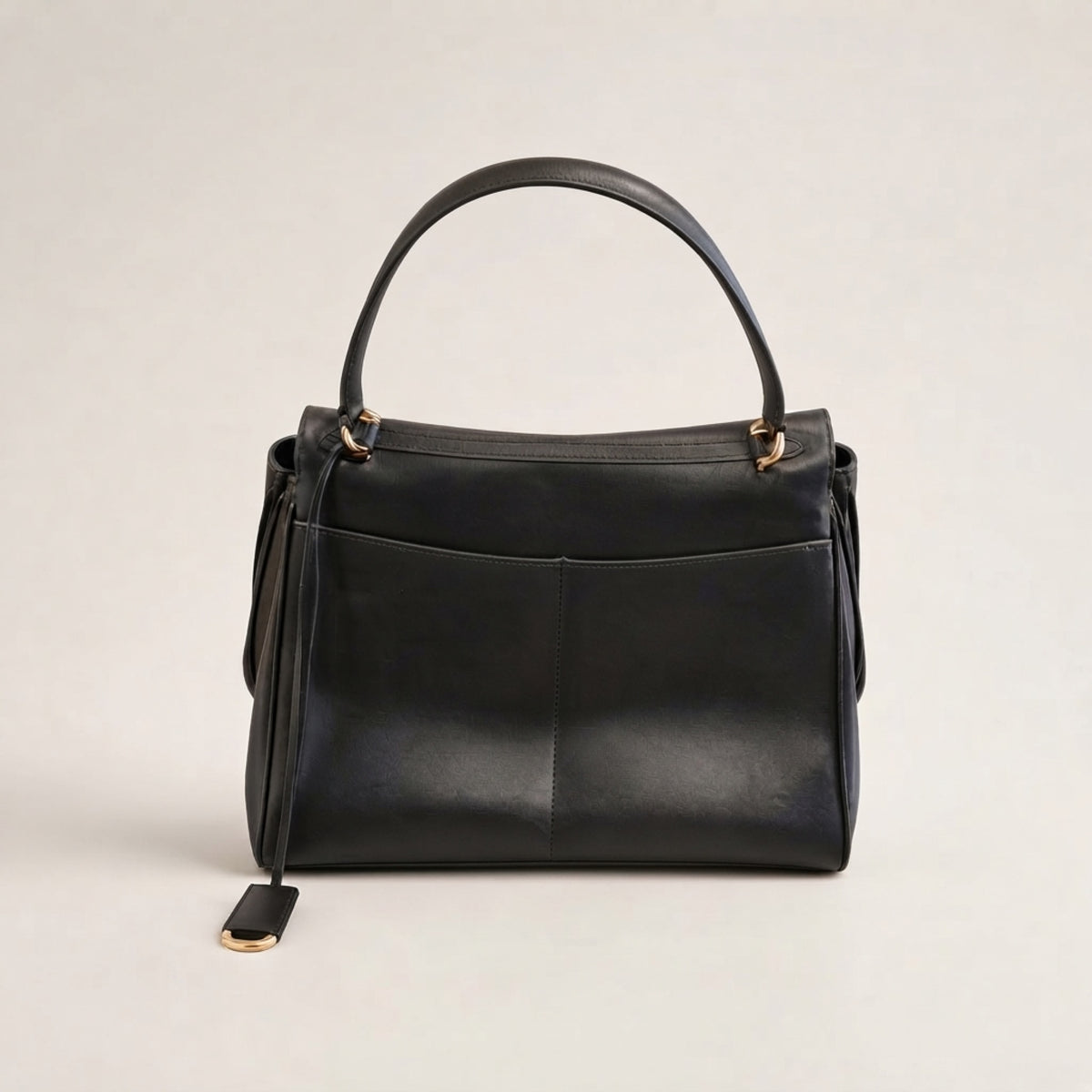 A black leather handbag with a top handle, gold-tone hardware, and a hanging leather tag, photographed against a neutral light gray background.