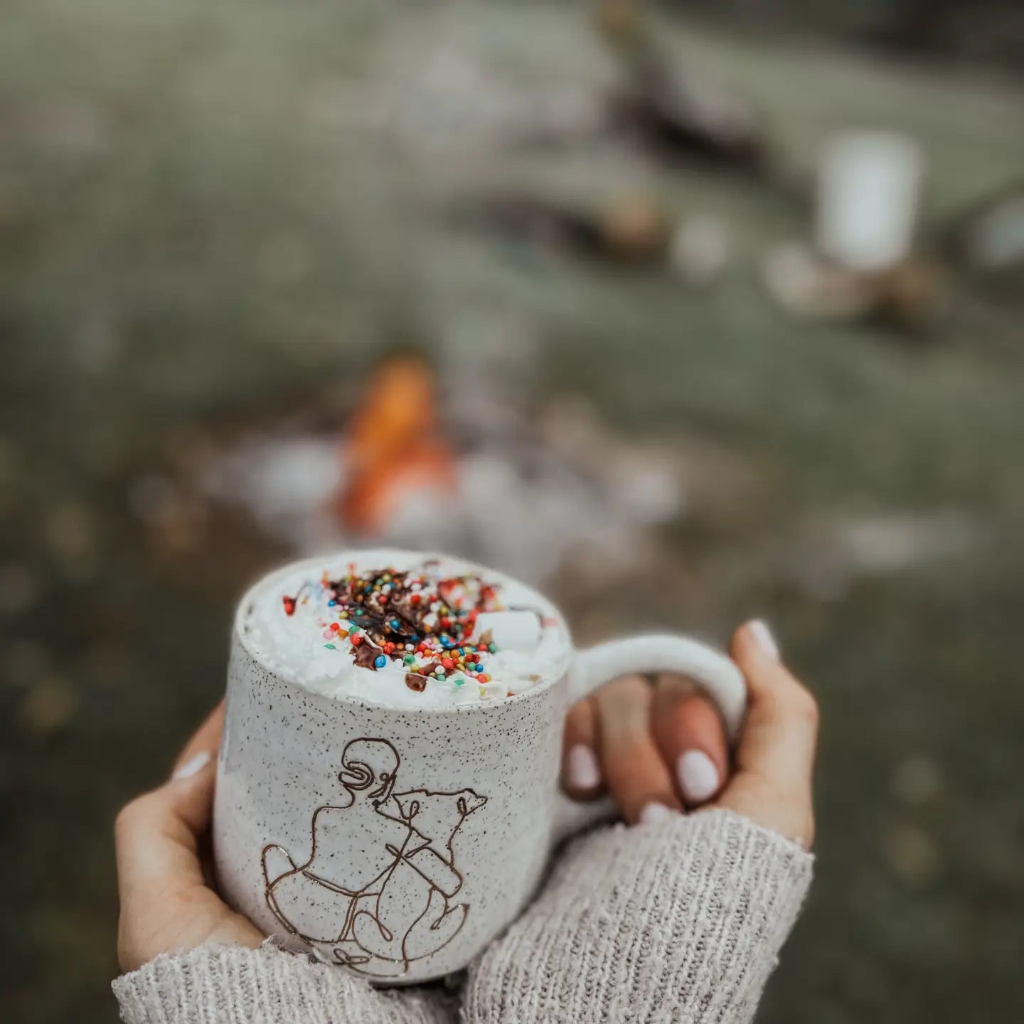 Hands holding a speckled ceramic mug with a gold girl-and-dog illustration, topped with whipped cream and sprinkles, in front of a blurred outdoor campfire